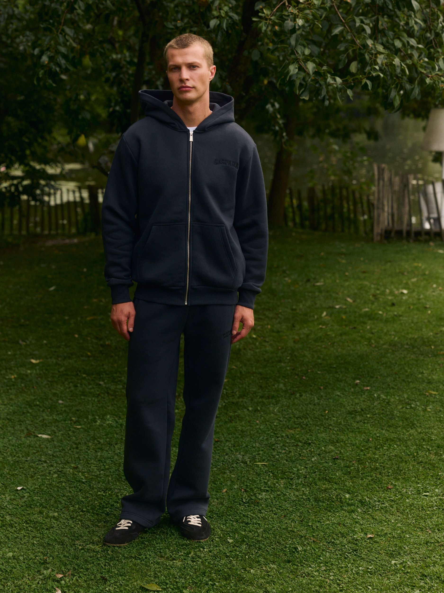 Man wearing a black zip-up hoodie and matching sweatpants standing outdoors on a lawn.