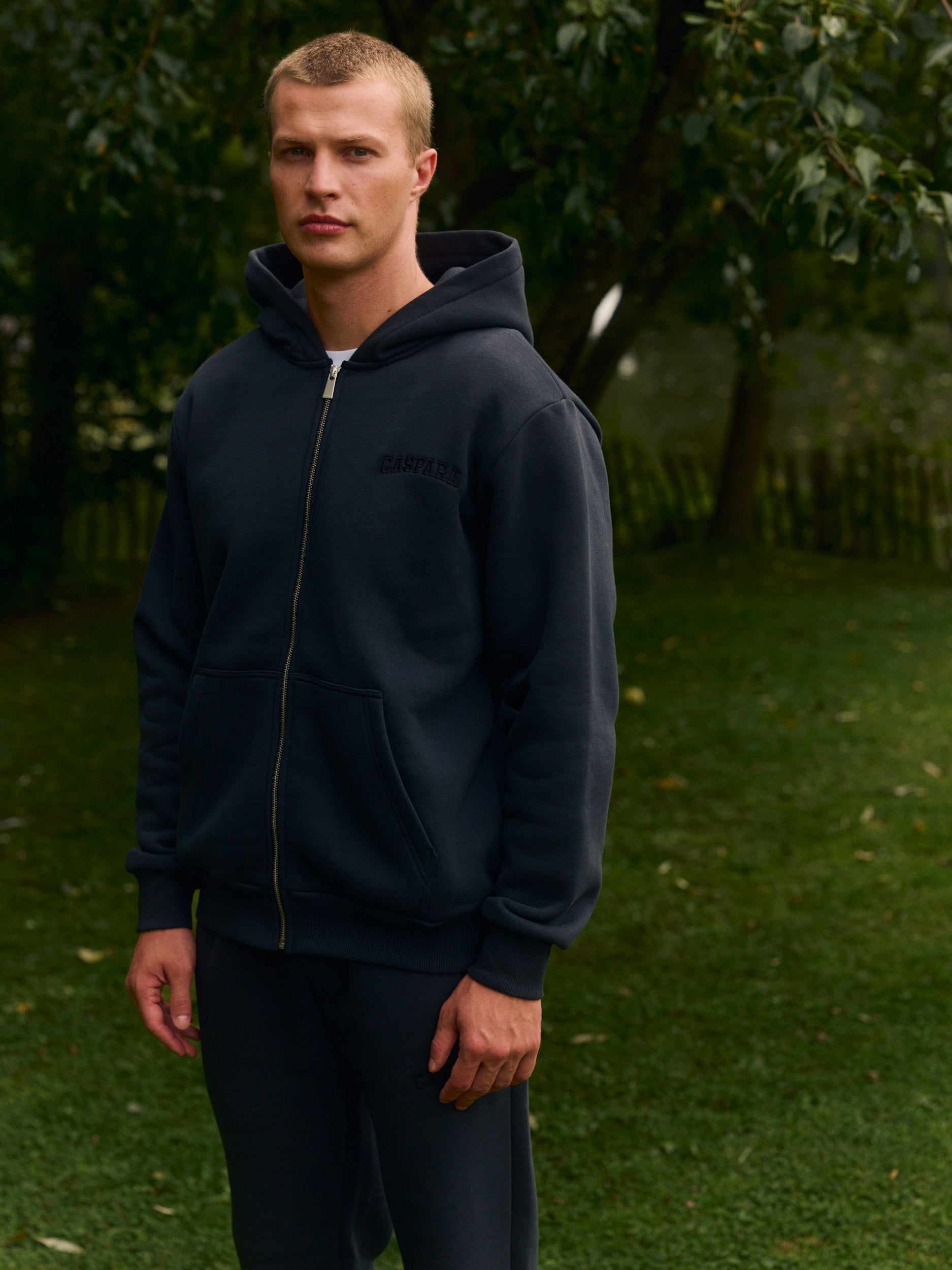 Man wearing a black zip-up hoodie standing outdoors in a lush green garden.