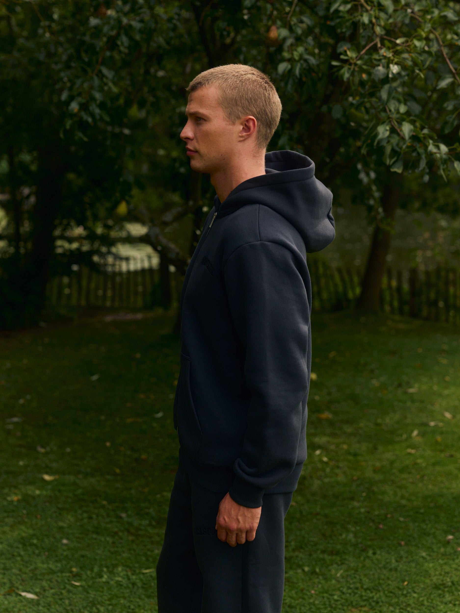 Man standing in profile wearing a black zip-up hoodie outdoors in a garden.