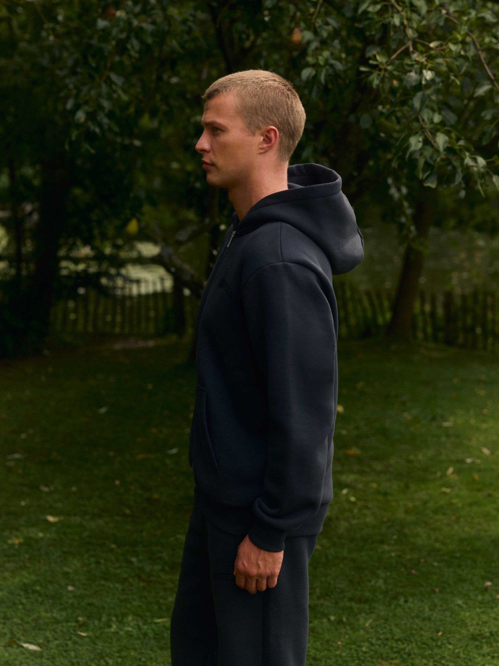 Man wearing a black CASPARA zip hoodie standing outdoors in a lush garden.