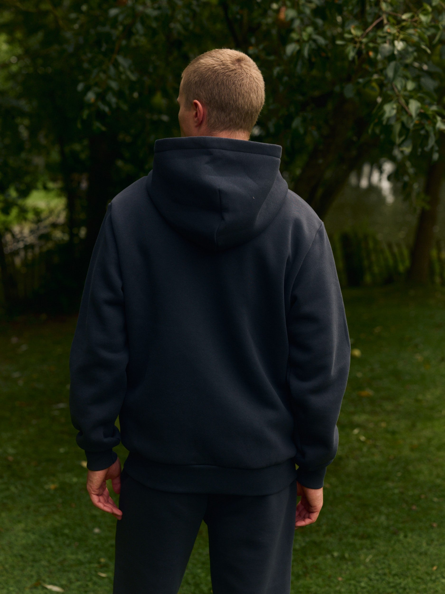 Rear view of a man wearing a black zip hoodie outdoors in front of green trees.