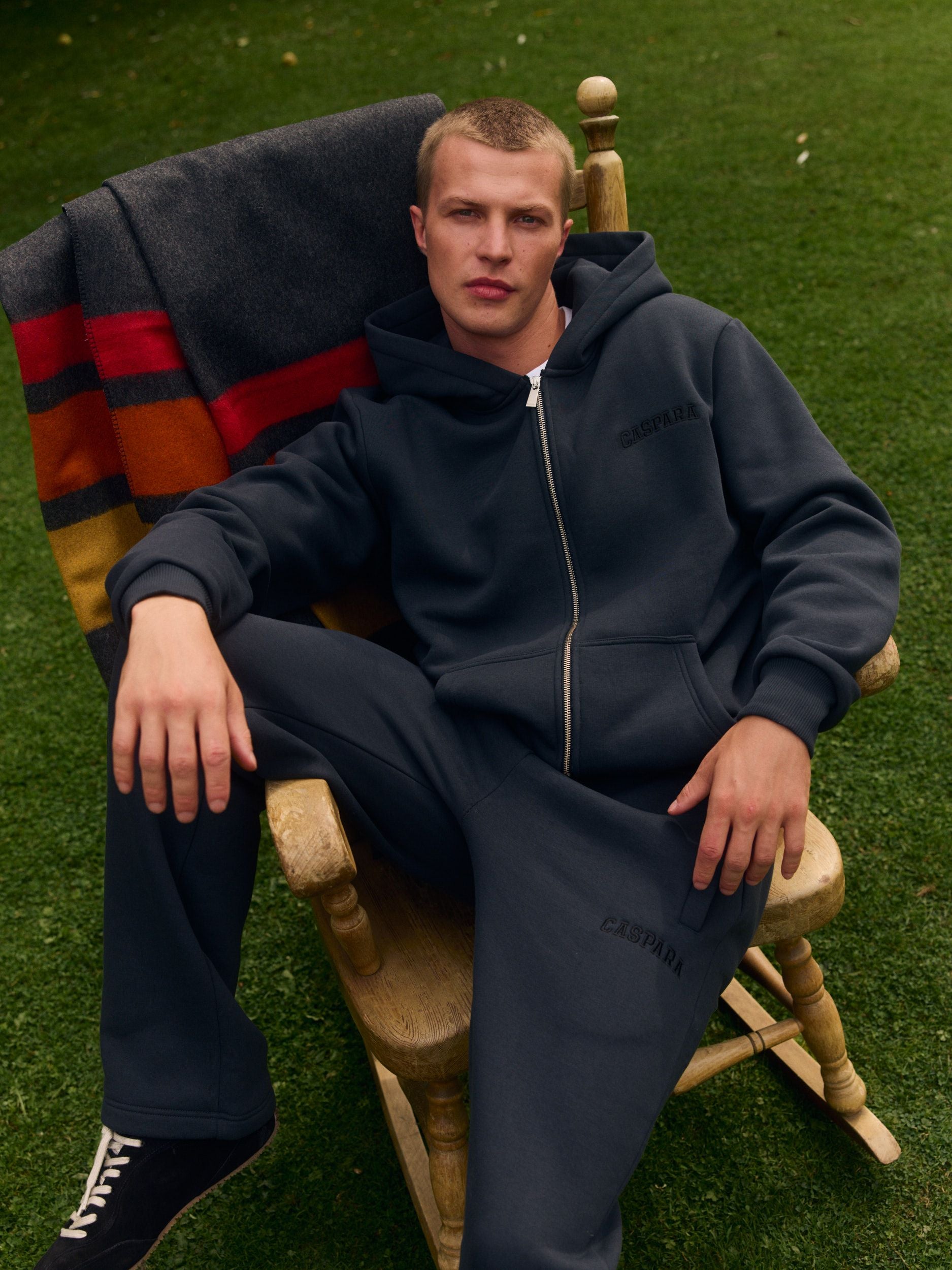 Man wearing a black zip hoodie and matching sweatpants sitting in a wooden rocking chair on grass.