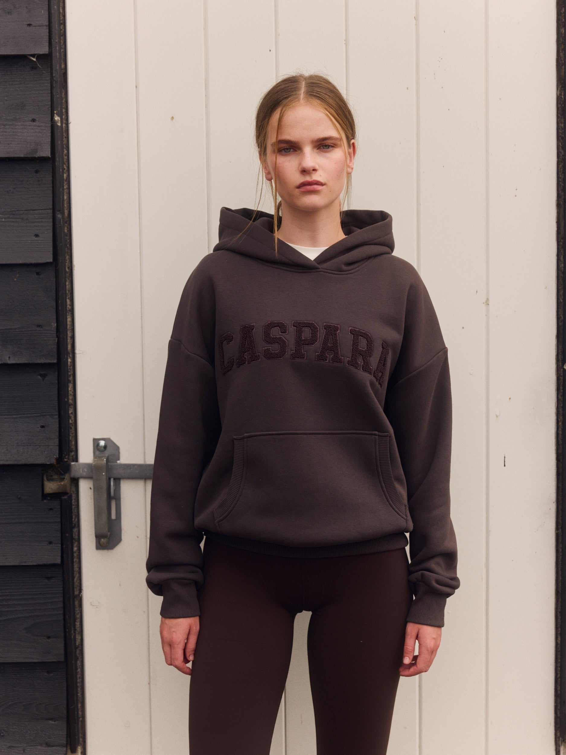 Woman wearing an espresso brown CASPARA hoodie and matching leggings in front of a white door.