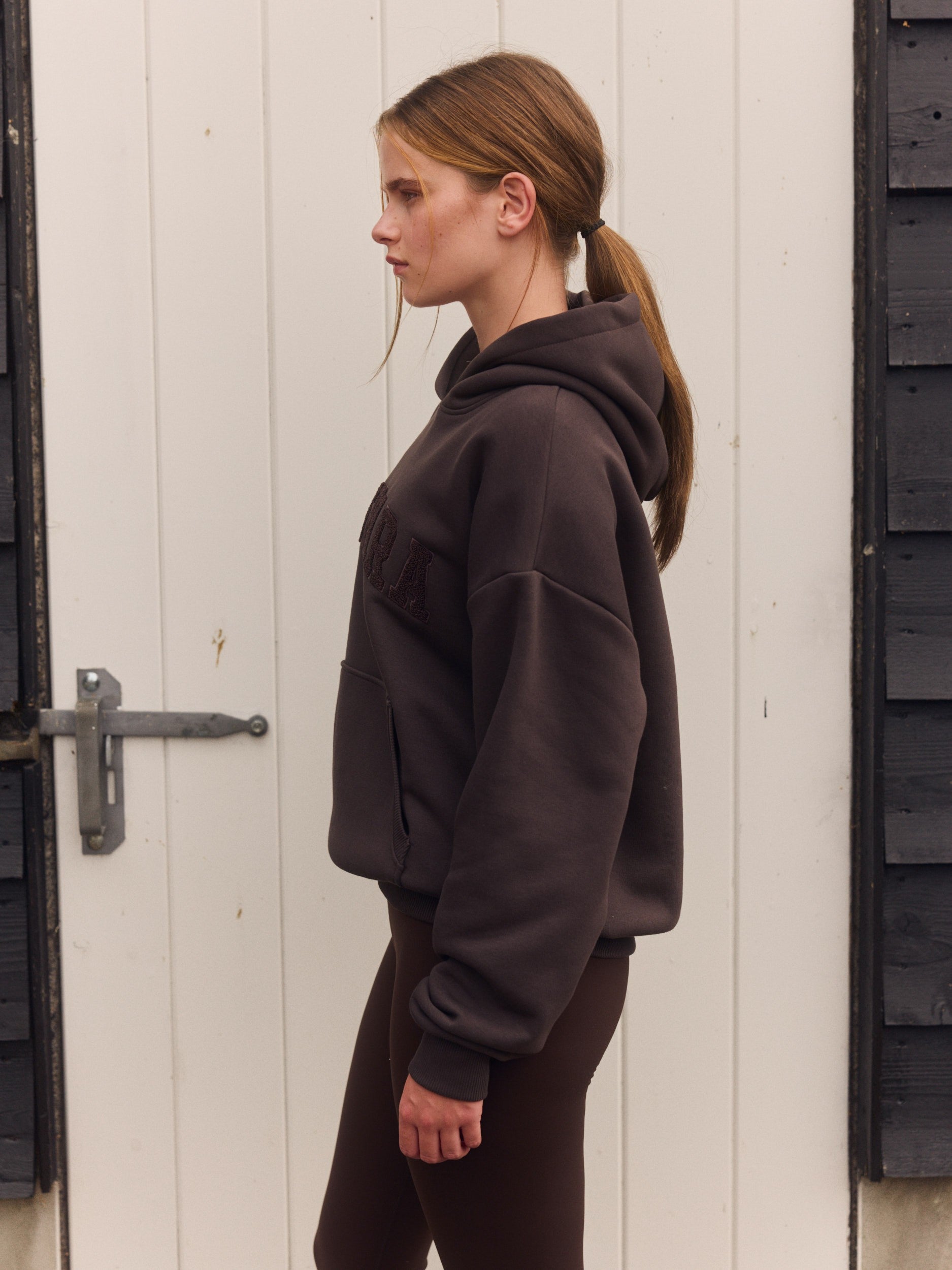 Woman wearing an espresso brown hoodie and matching leggings in profile against a white wooden door.