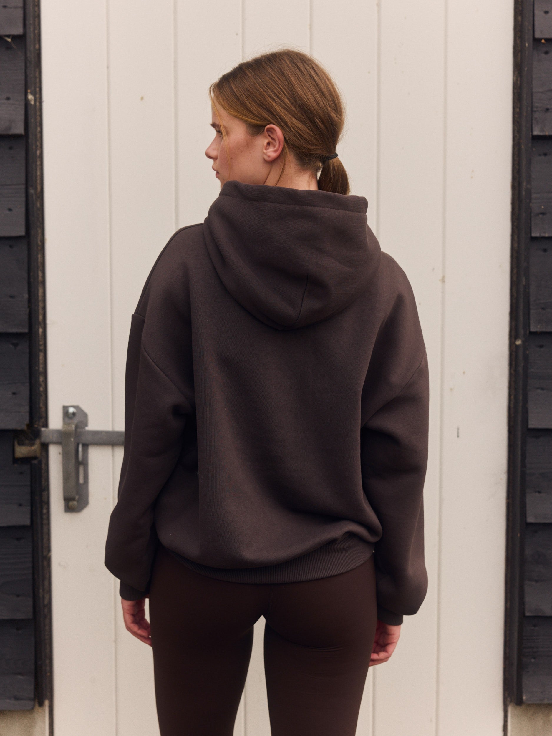 Woman from behind wearing an espresso brown zip hoodie and matching leggings in front of a white door.