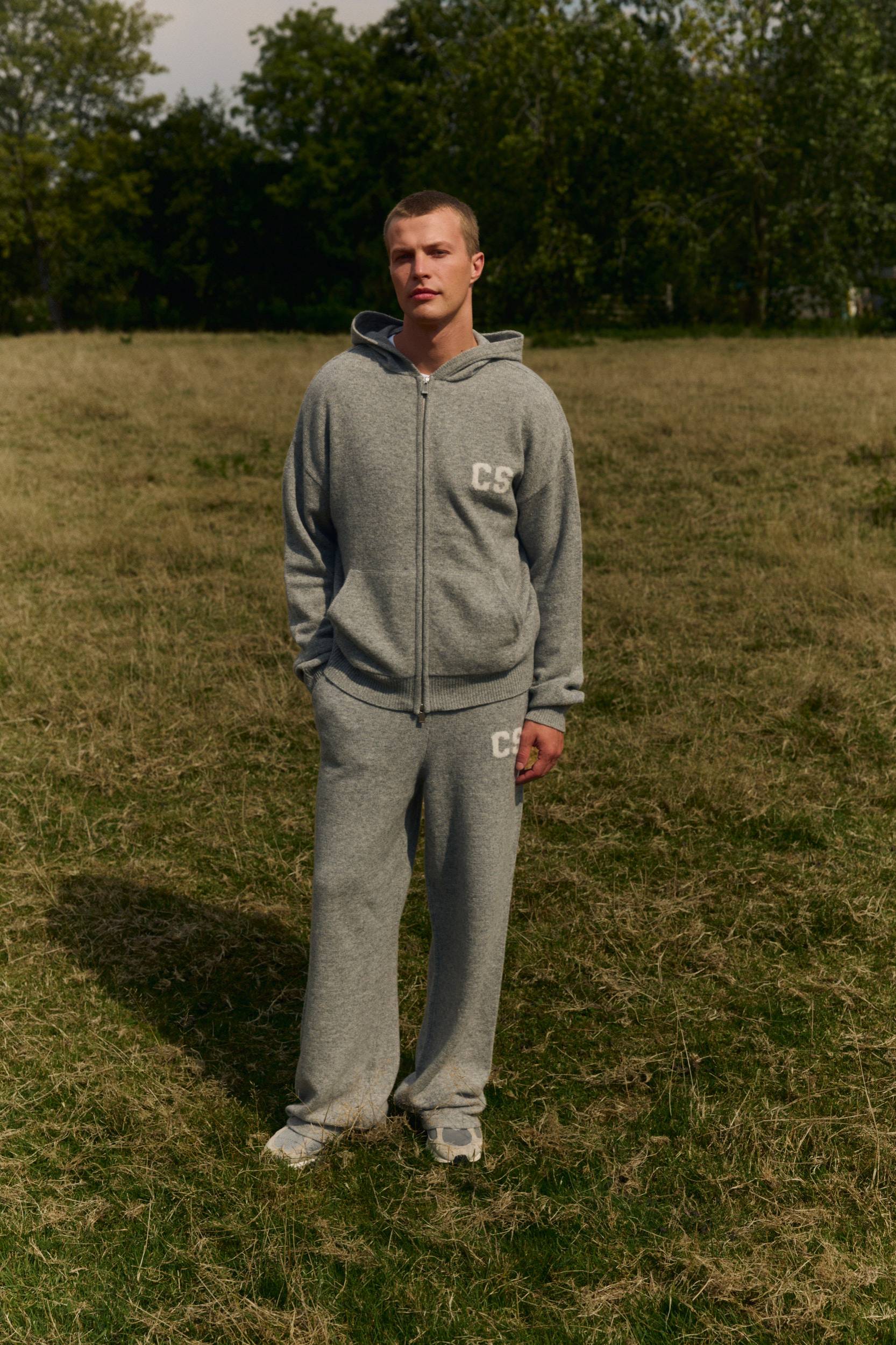 Man wearing dark grey melange woolknit straight-leg pants and a matching hoodie in a field.
