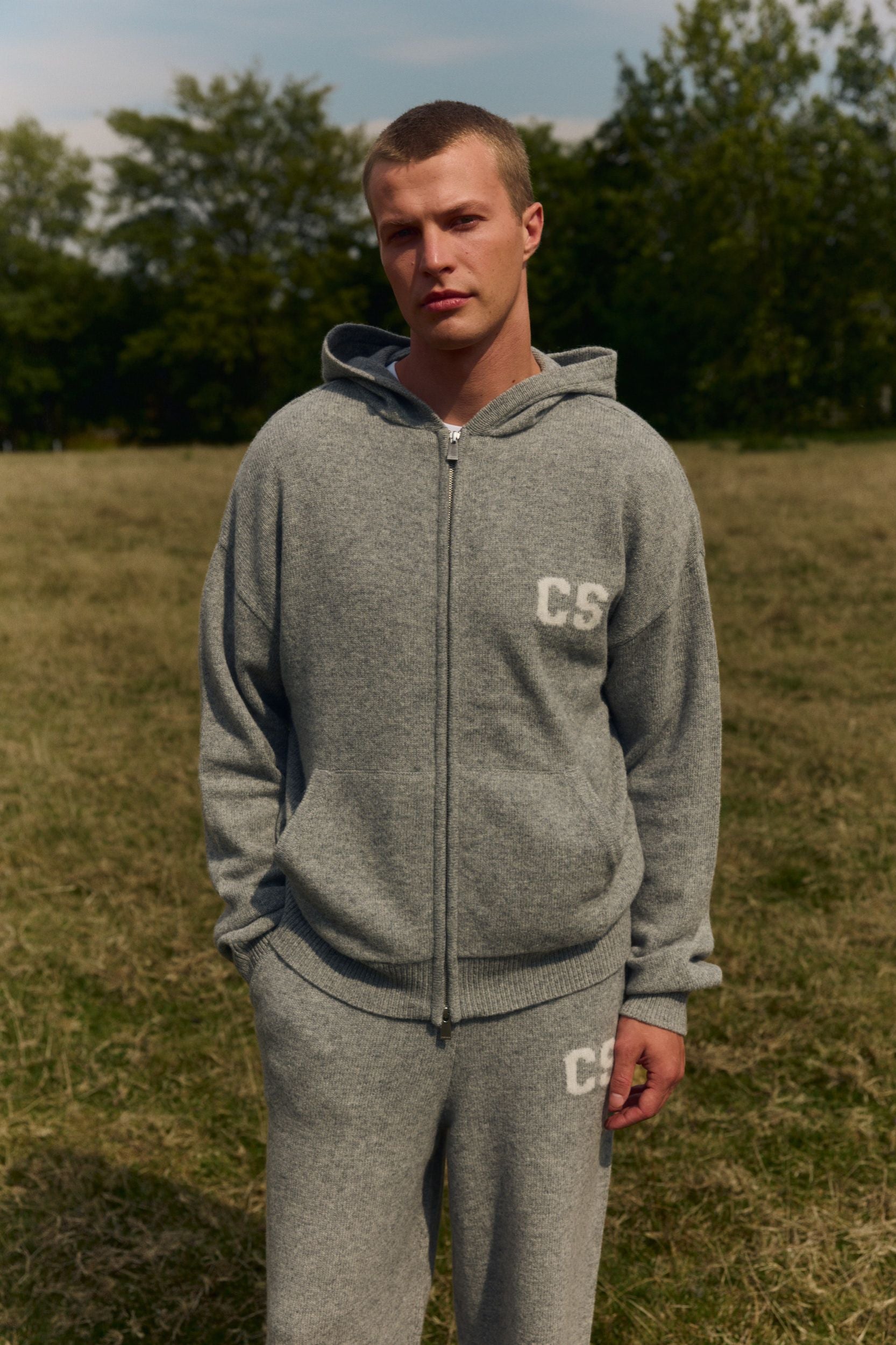 Man wearing a dark grey wool knit zip hoodie standing in a grassy field.