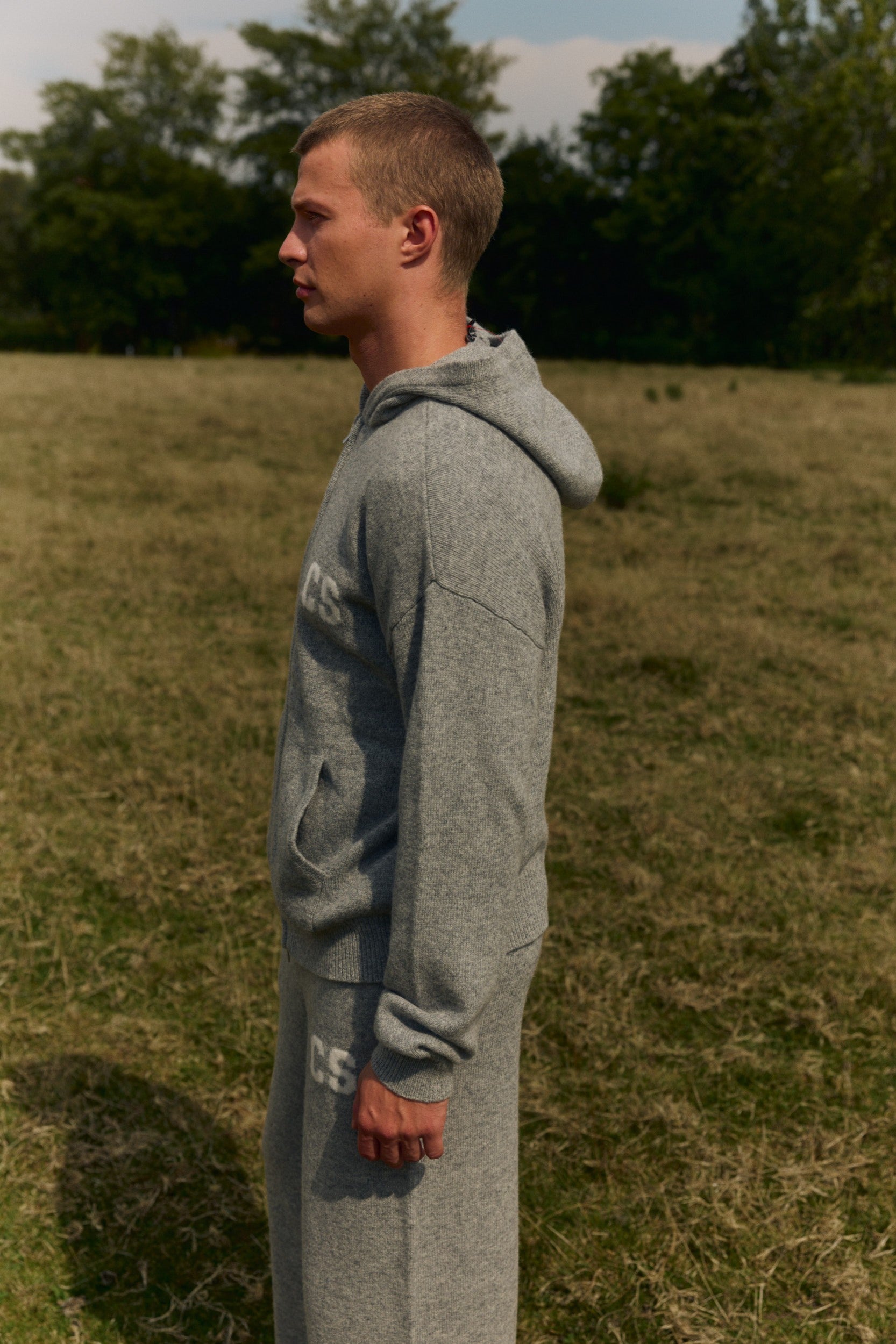 Man wearing a grey wool-knit zip hoodie standing outdoors in a grassy field.