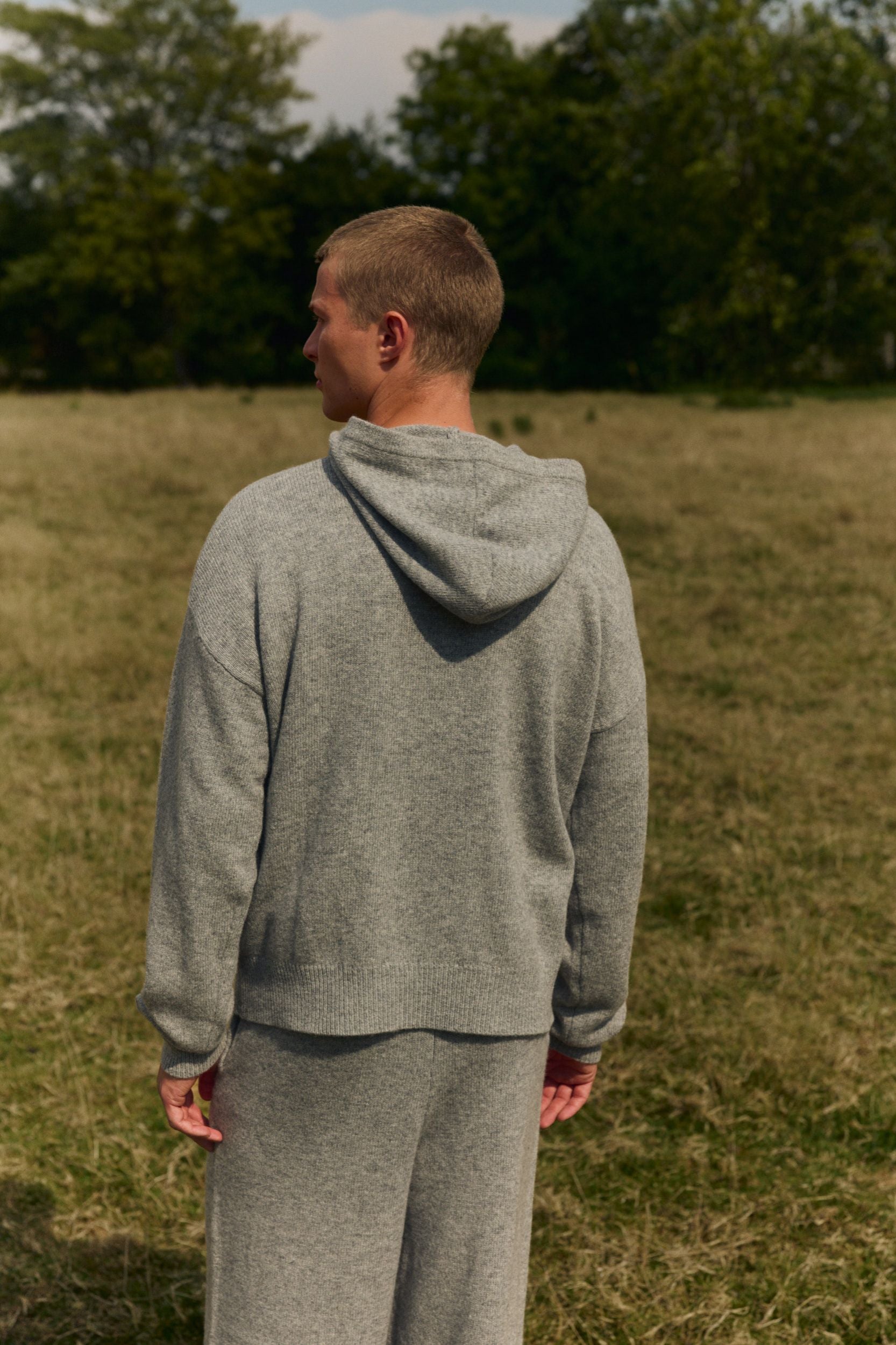 Back view of a man wearing a grey melange woolknit hooded jacket in an open field.