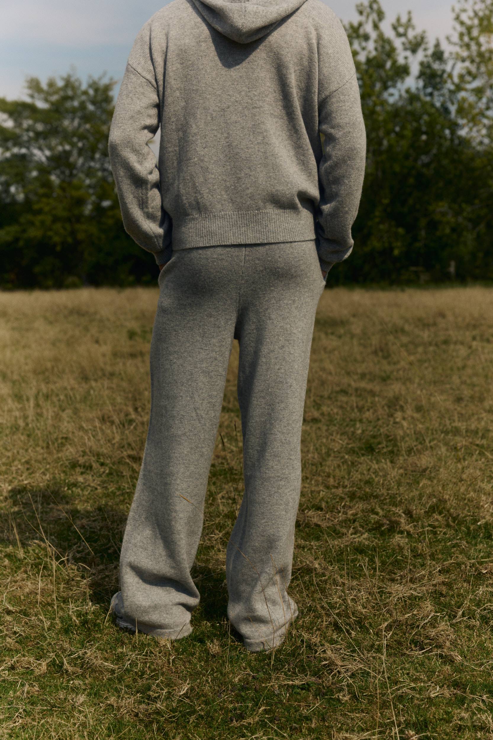 Rear view of a man wearing dark grey melange wool knit pants and a matching hoodie in a grassy field.
