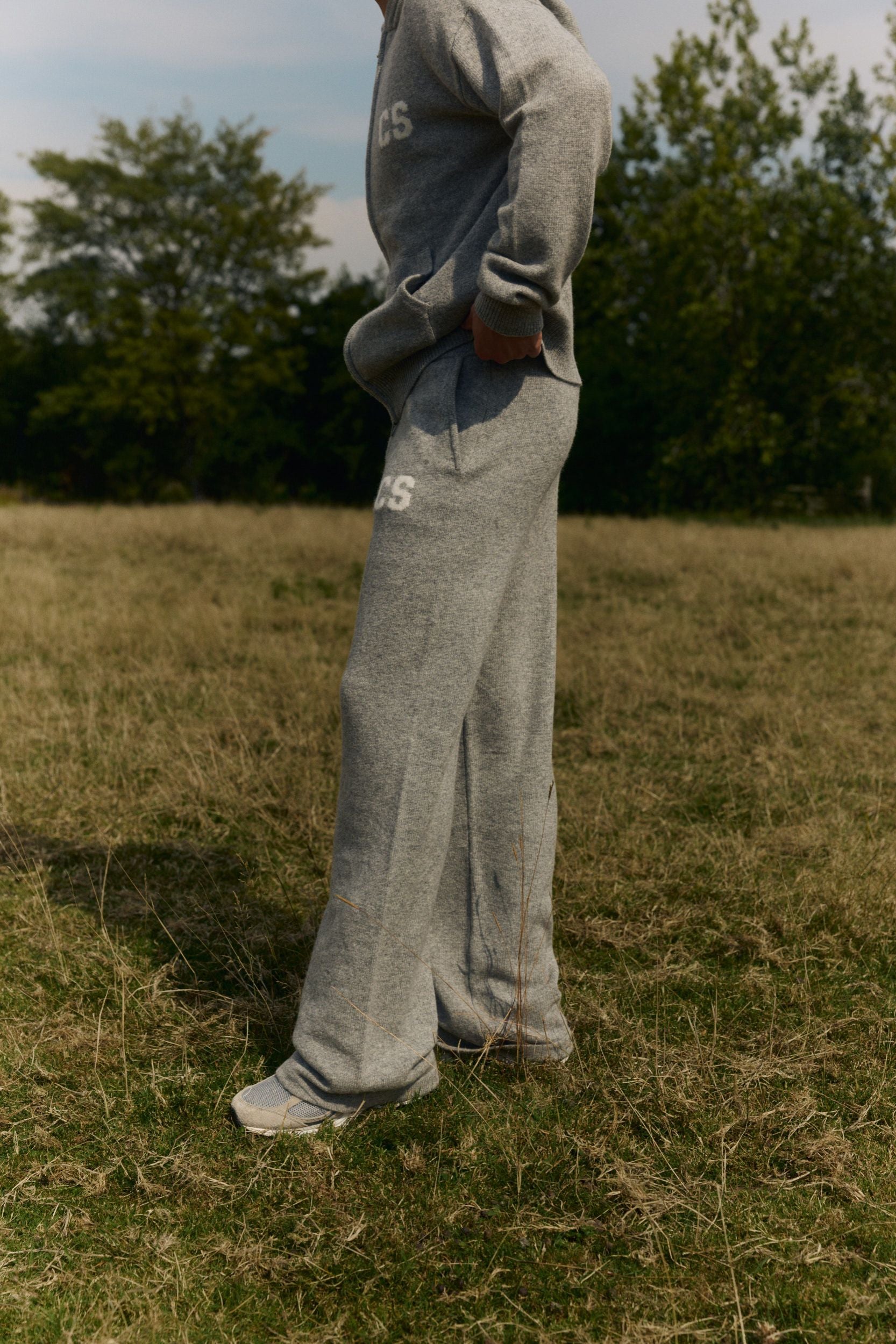 Man wearing dark grey melange woolknit straight-leg pants and matching hoodie in a grassy field.