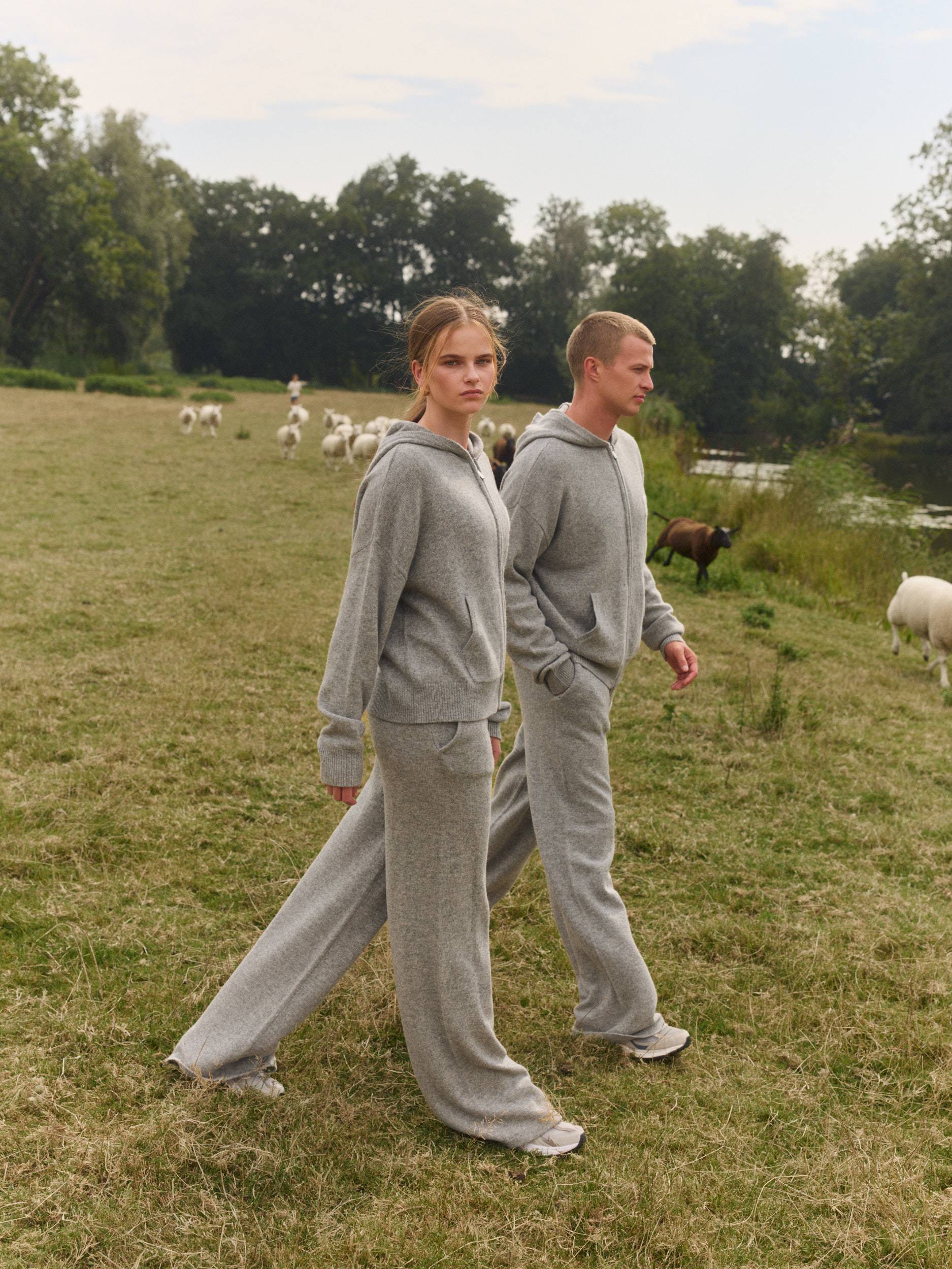 Man and woman wearing matching grey woolknit hoodies and straight-leg pants walking in a field.