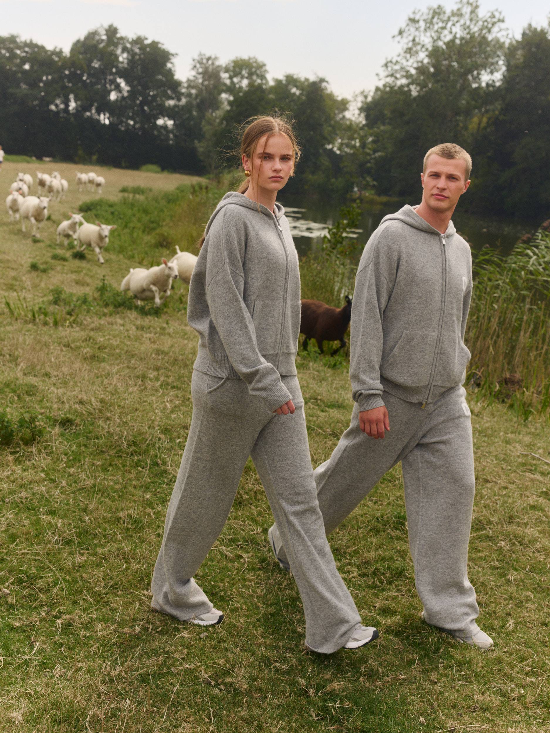 Man and woman wearing matching grey knit zip hoodies and straight-leg pants in a grassy field.