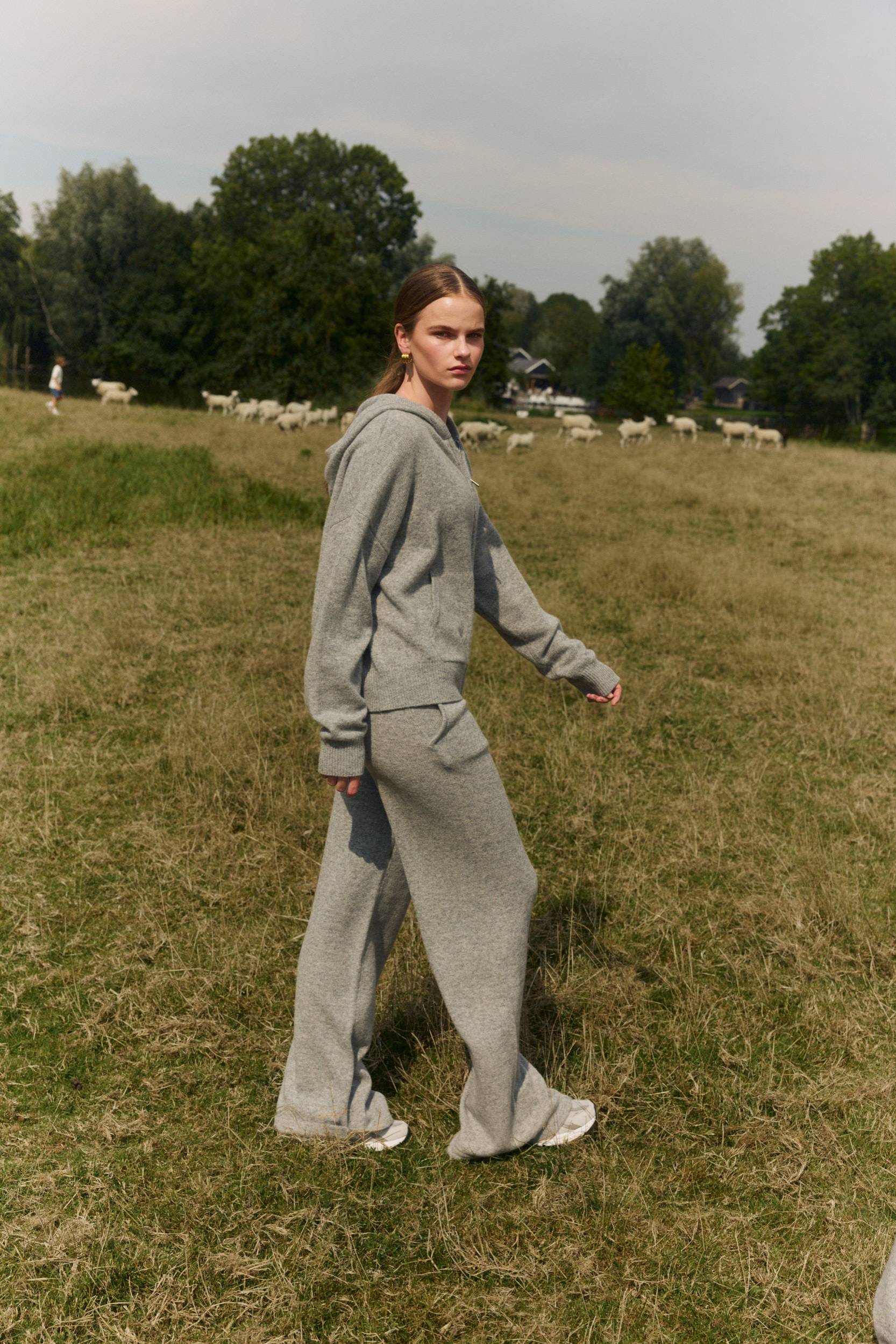 Woman wearing grey knit straight-leg pants and a matching hoodie in a grassy field.