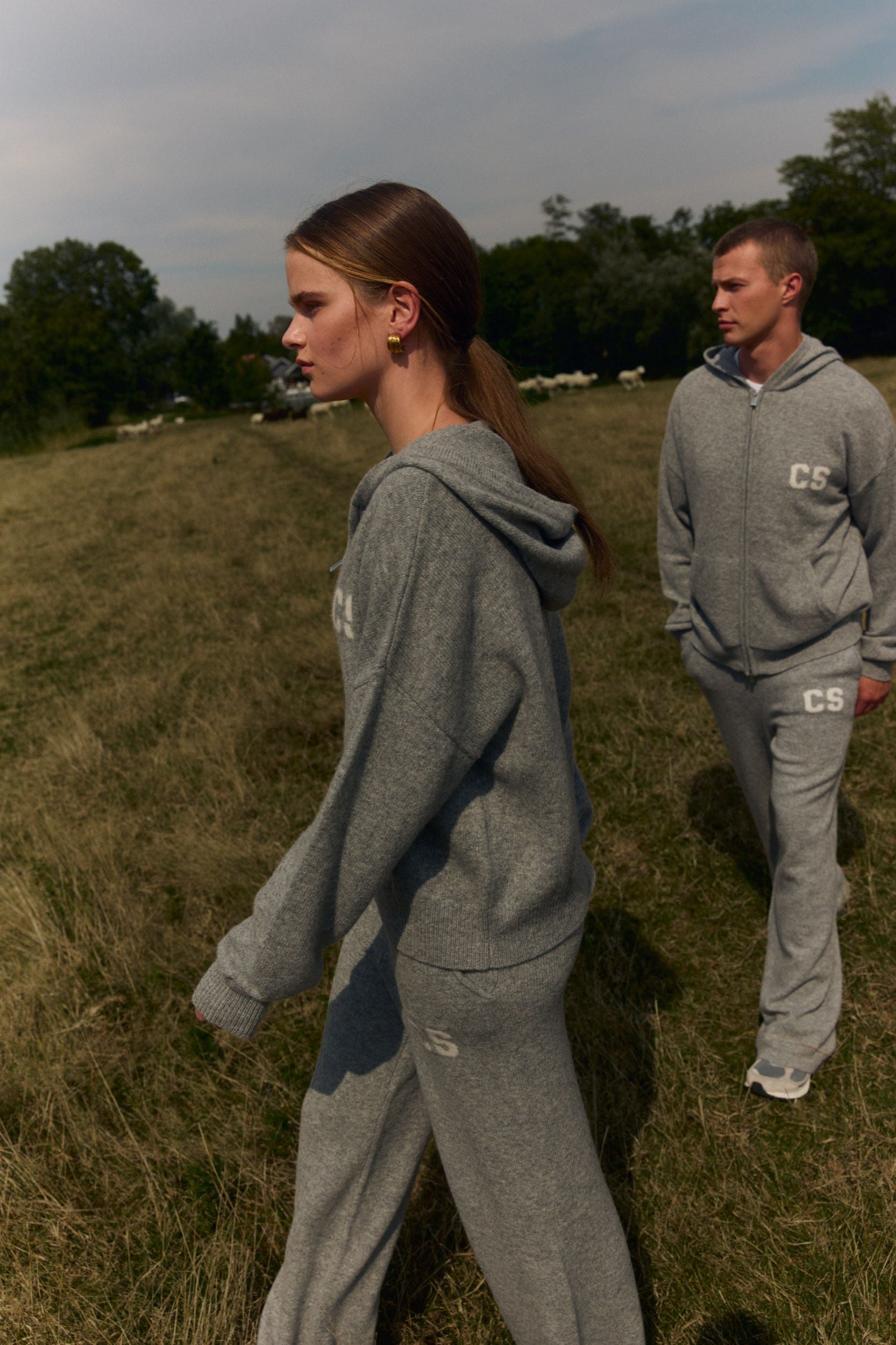 Woman wearing grey woolknit straight-leg pants and matching hoodie walking through a grassy field.