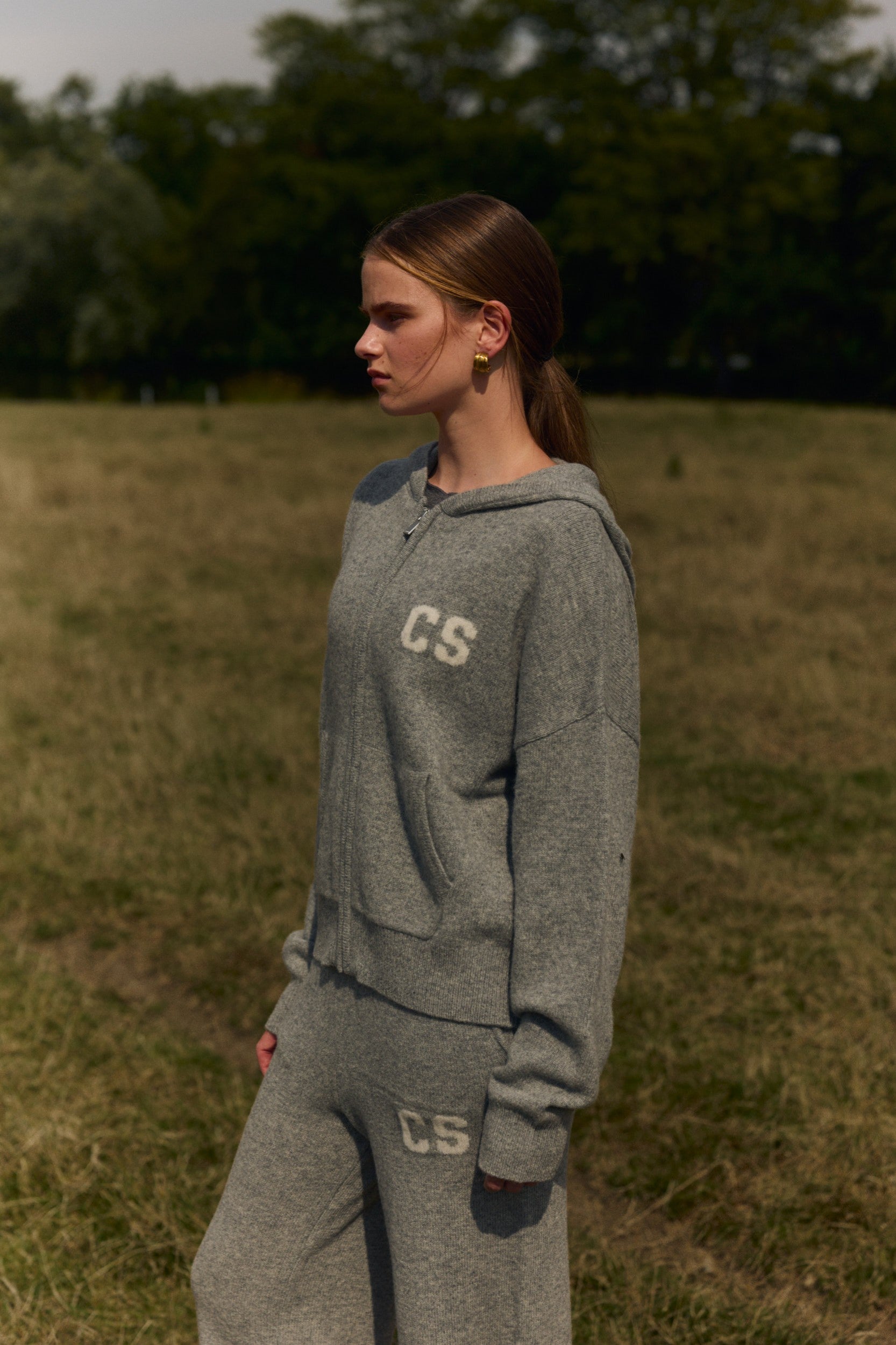 Woman wearing a grey knit zip jacket with CS logo standing in a grassy field.