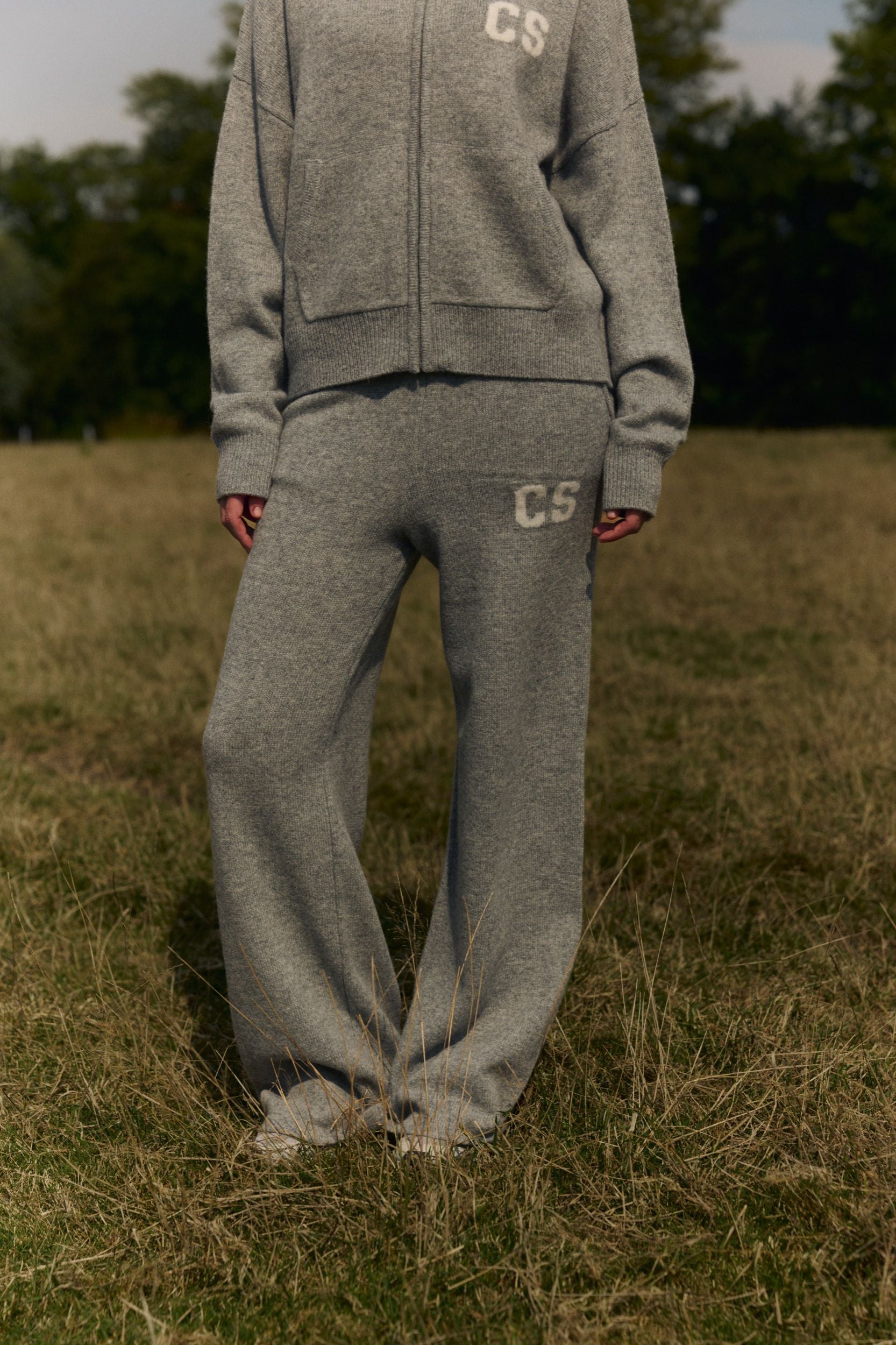 Woman wearing grey knit straight-leg pants and a matching zip-up hoodie in a grassy field.