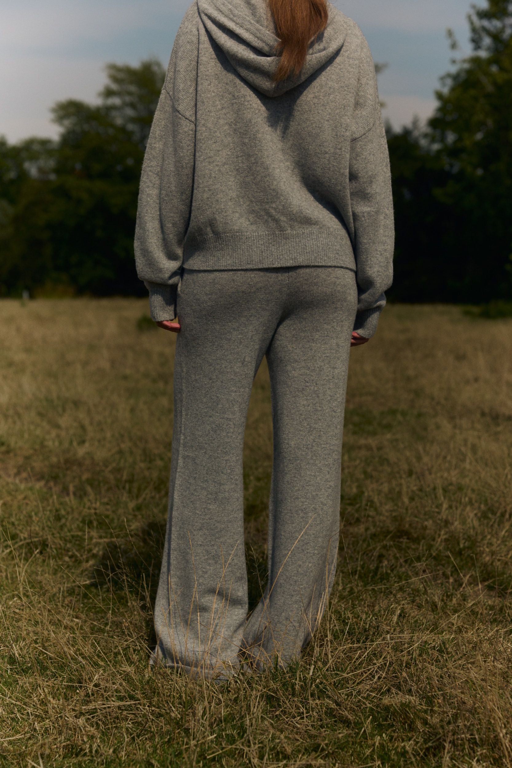 Back view of a woman wearing grey knit straight-leg pants and a matching hoodie in an open field.