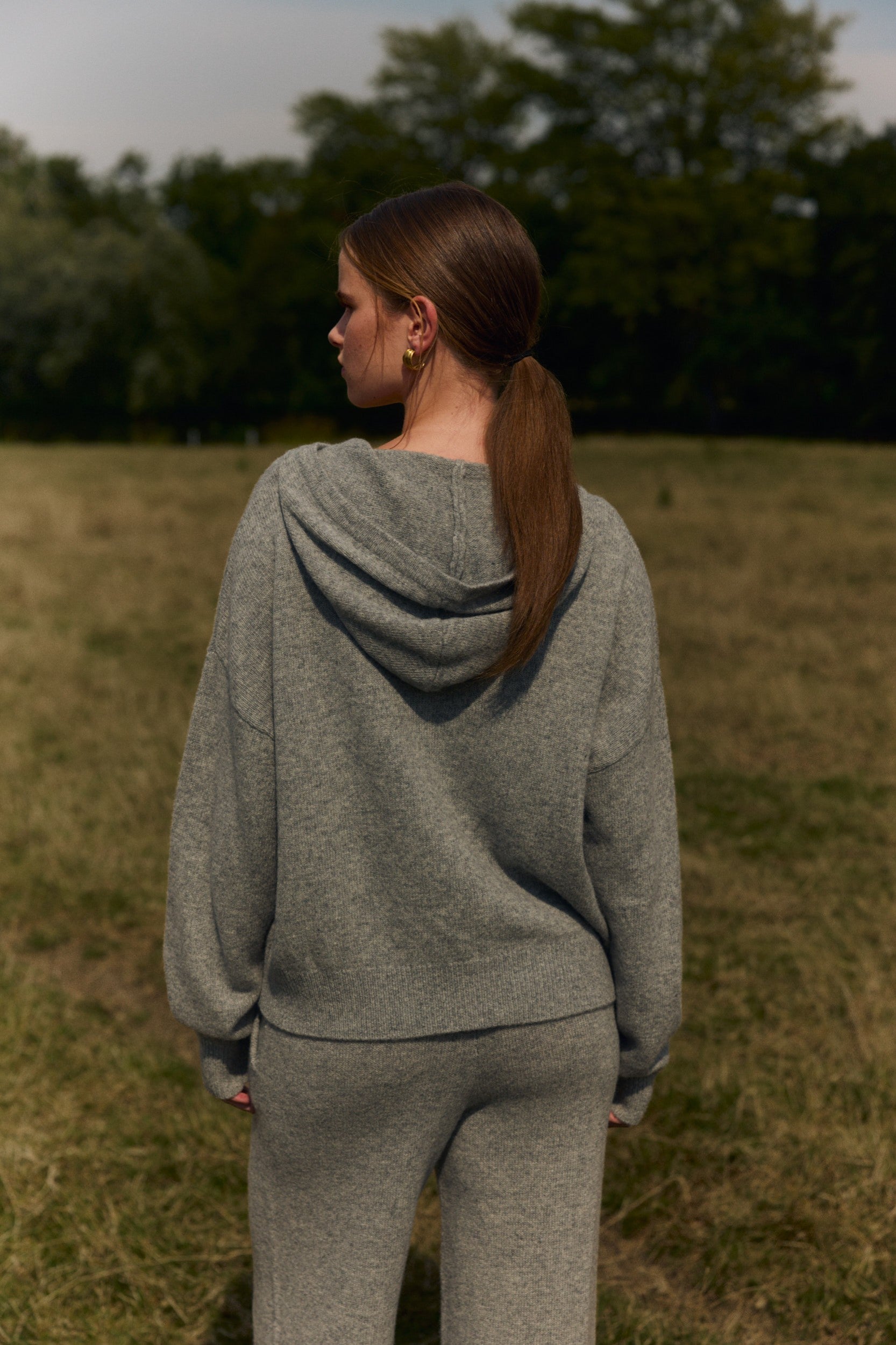 Woman seen from behind wearing a grey knit hooded jacket and matching pants in a grassy field.