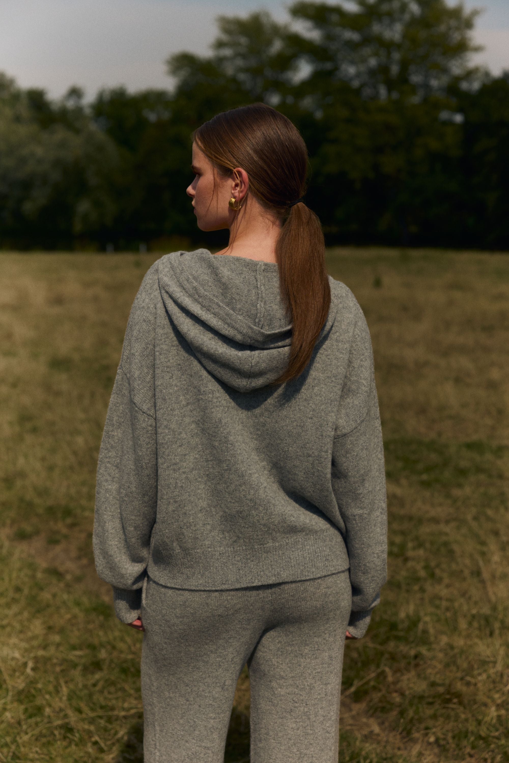 Back view of a woman wearing a grey CASPARA woolknit zip jacket outdoors in an open grassy field.