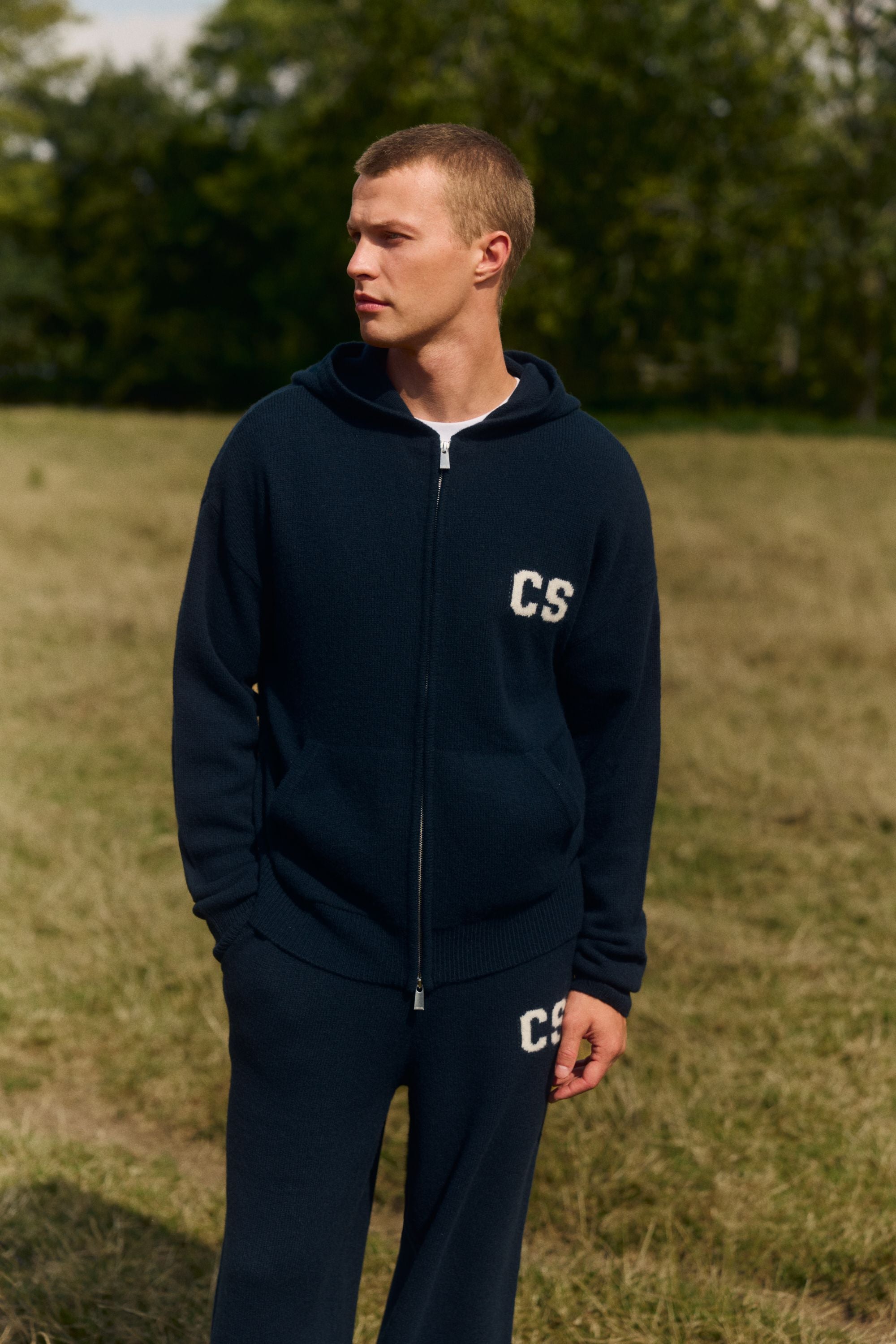 Man wearing a navy CASPARA wool knit zip-up hoodie and matching pants standing outdoors in a field.