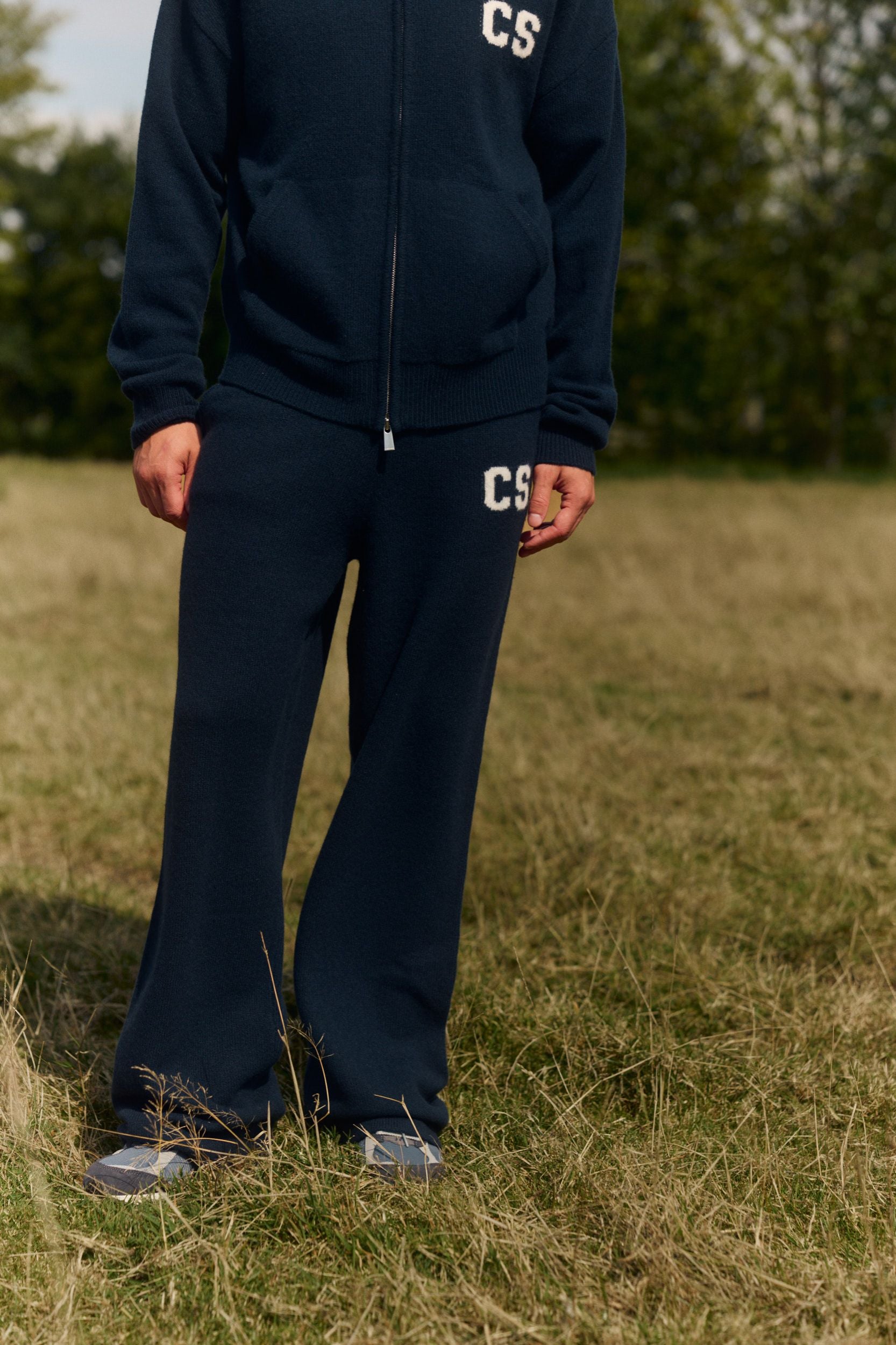 Man wearing navy wool knit straight-leg pants and a matching zip sweater in a grassy field.