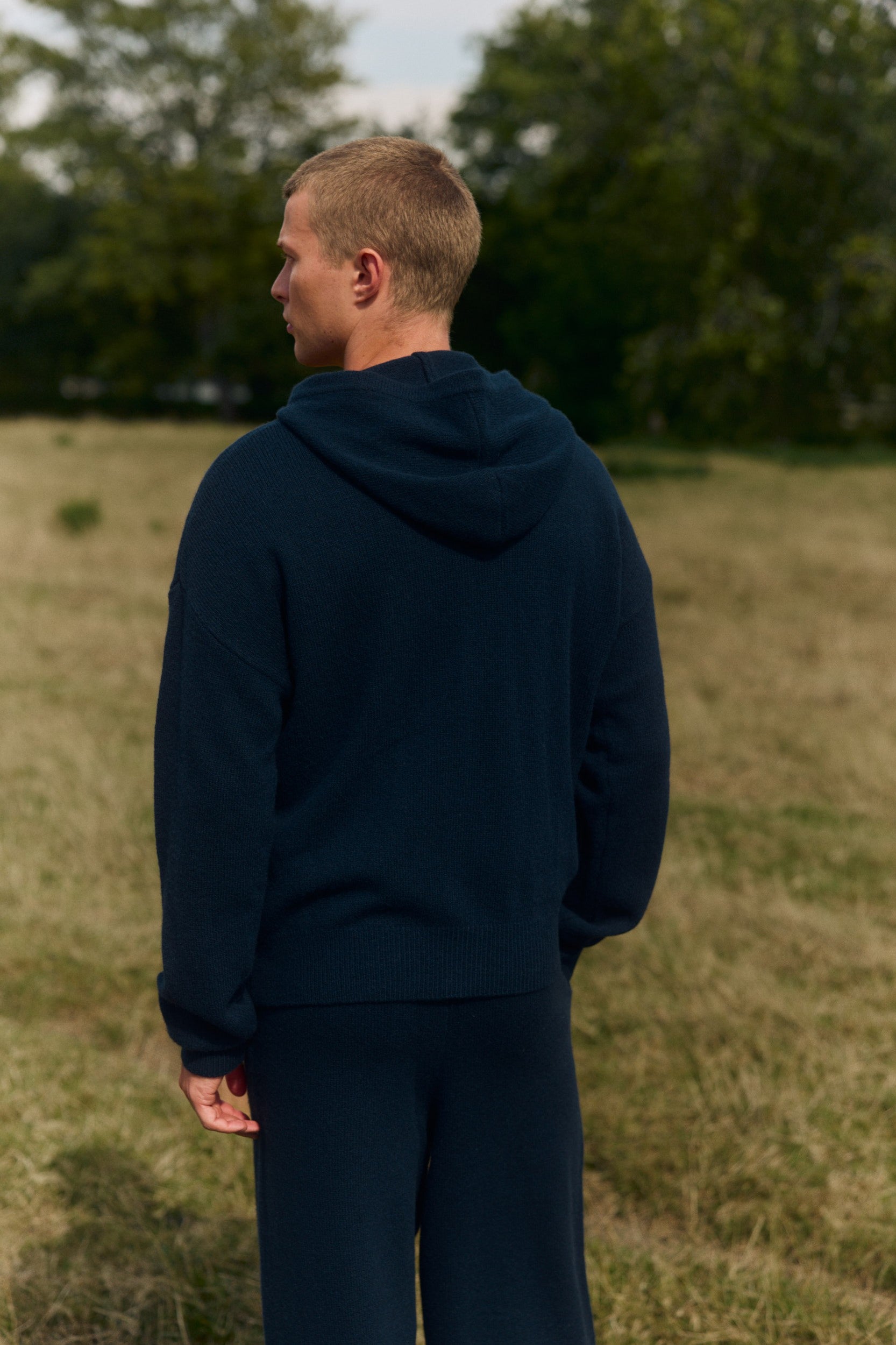 Man wearing a navy wool knit hooded jacket standing outdoors in a grassy field, seen from behind.