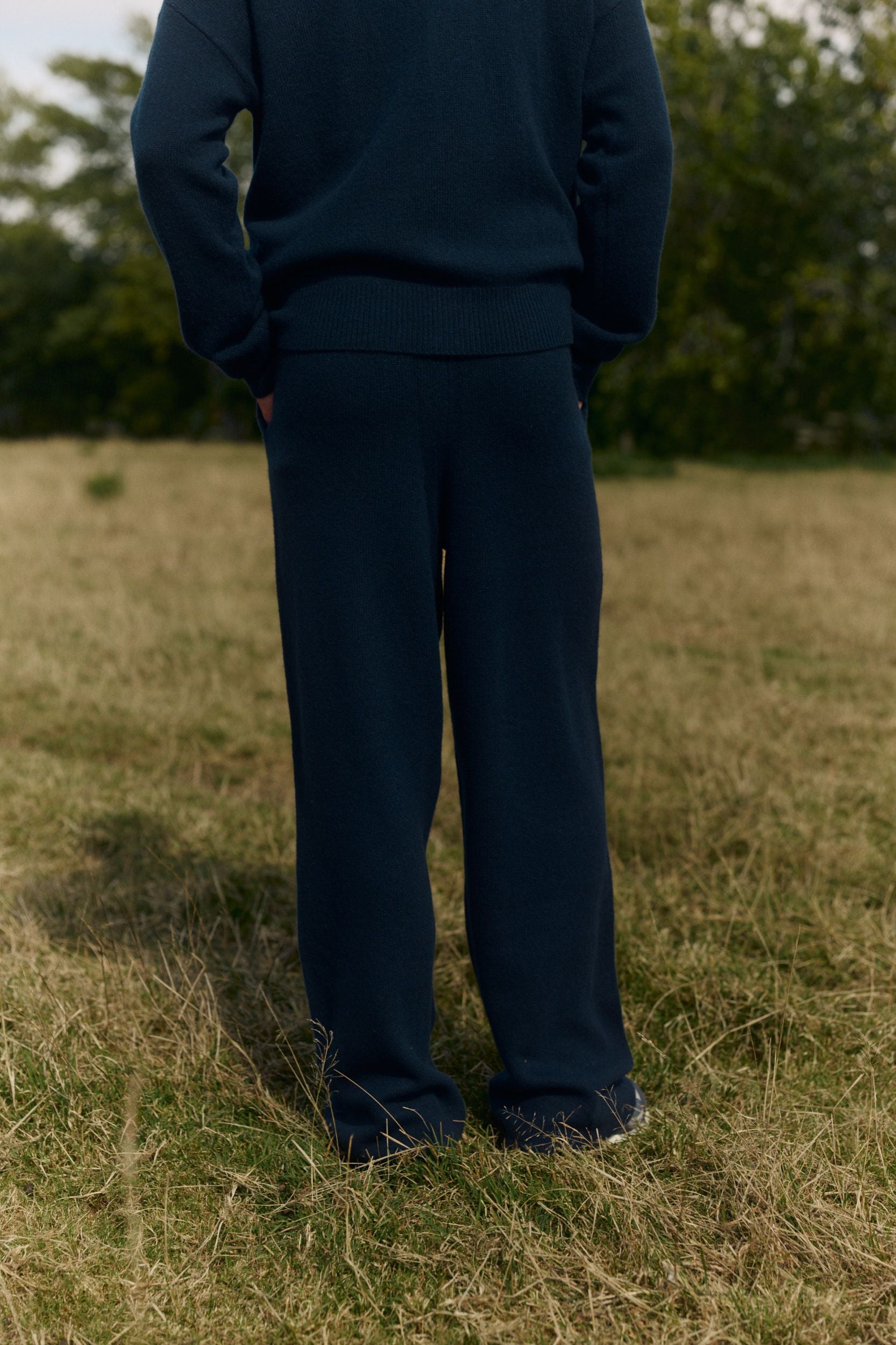 Rear view of a man wearing navy wool knit straight-leg pants and a matching sweater in a grassy field.