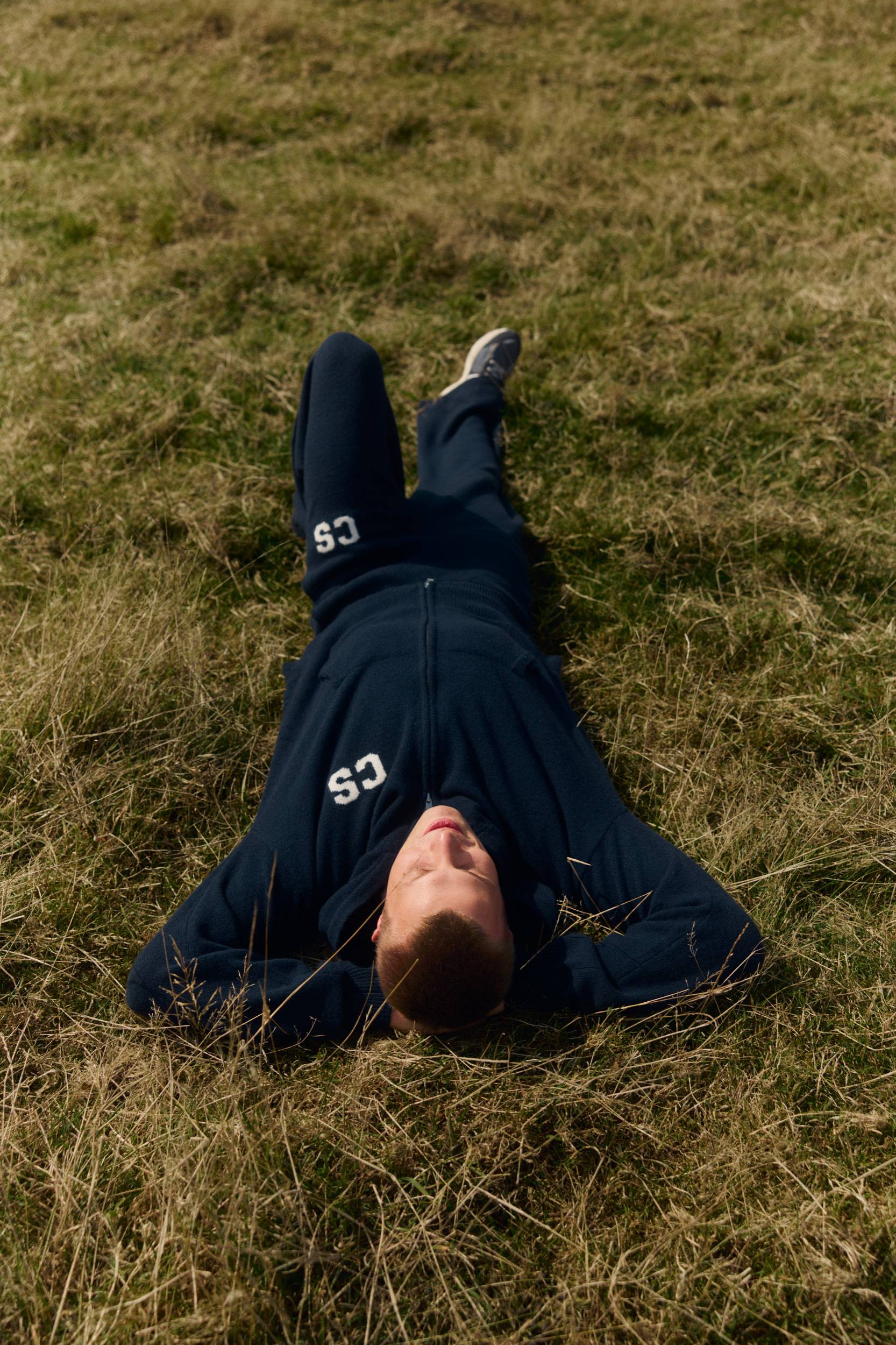 Man lying on a grassy field wearing a navy woolknit tracksuit with CS embroidery.