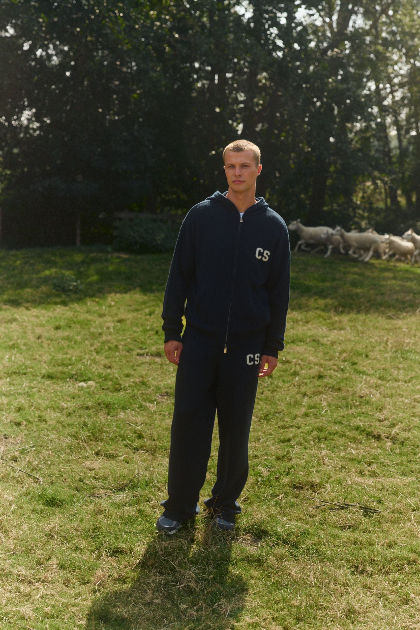 Man wearing a navy wool knit zip jacket and matching pants in a grassy field with sheep.