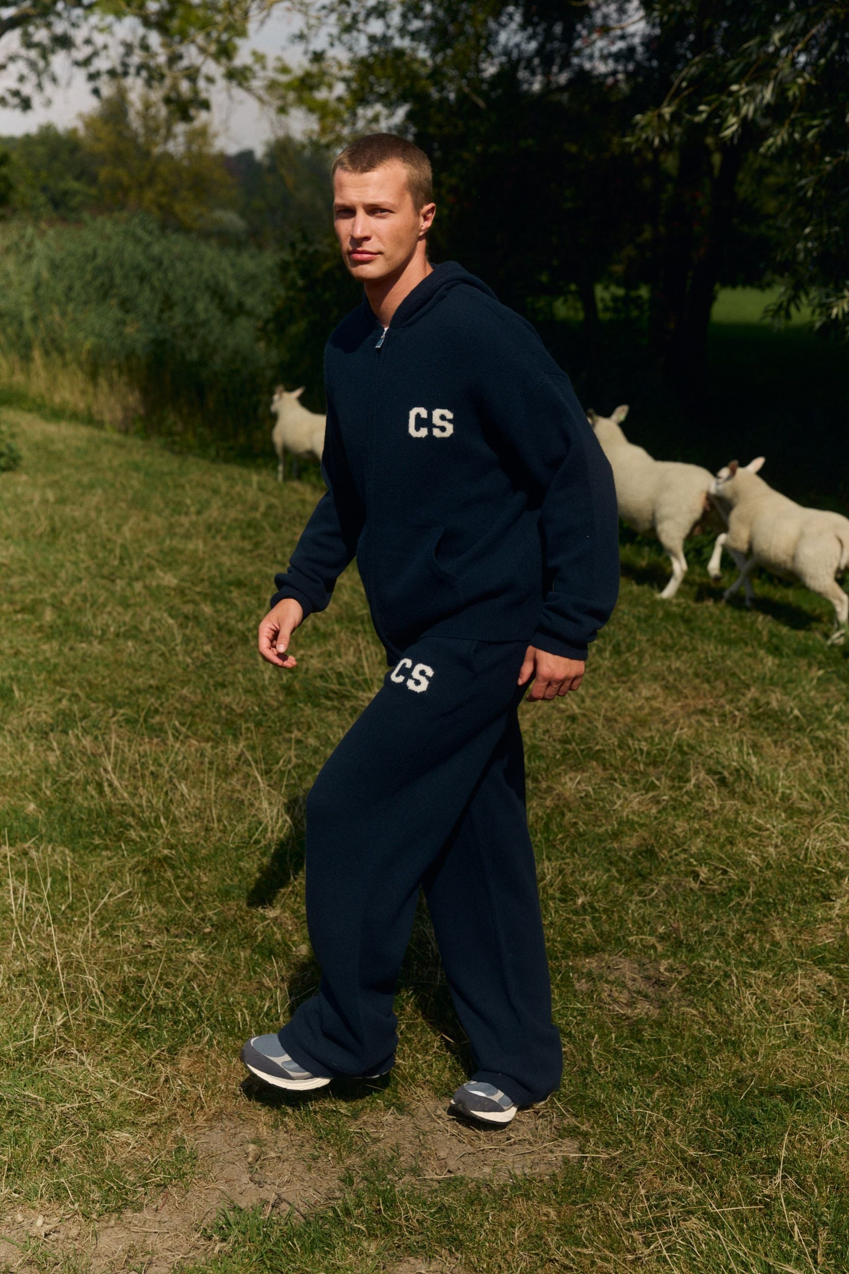 Man wearing navy woolknit straight leg pants and a matching hoodie walking in a grassy field with sheep.