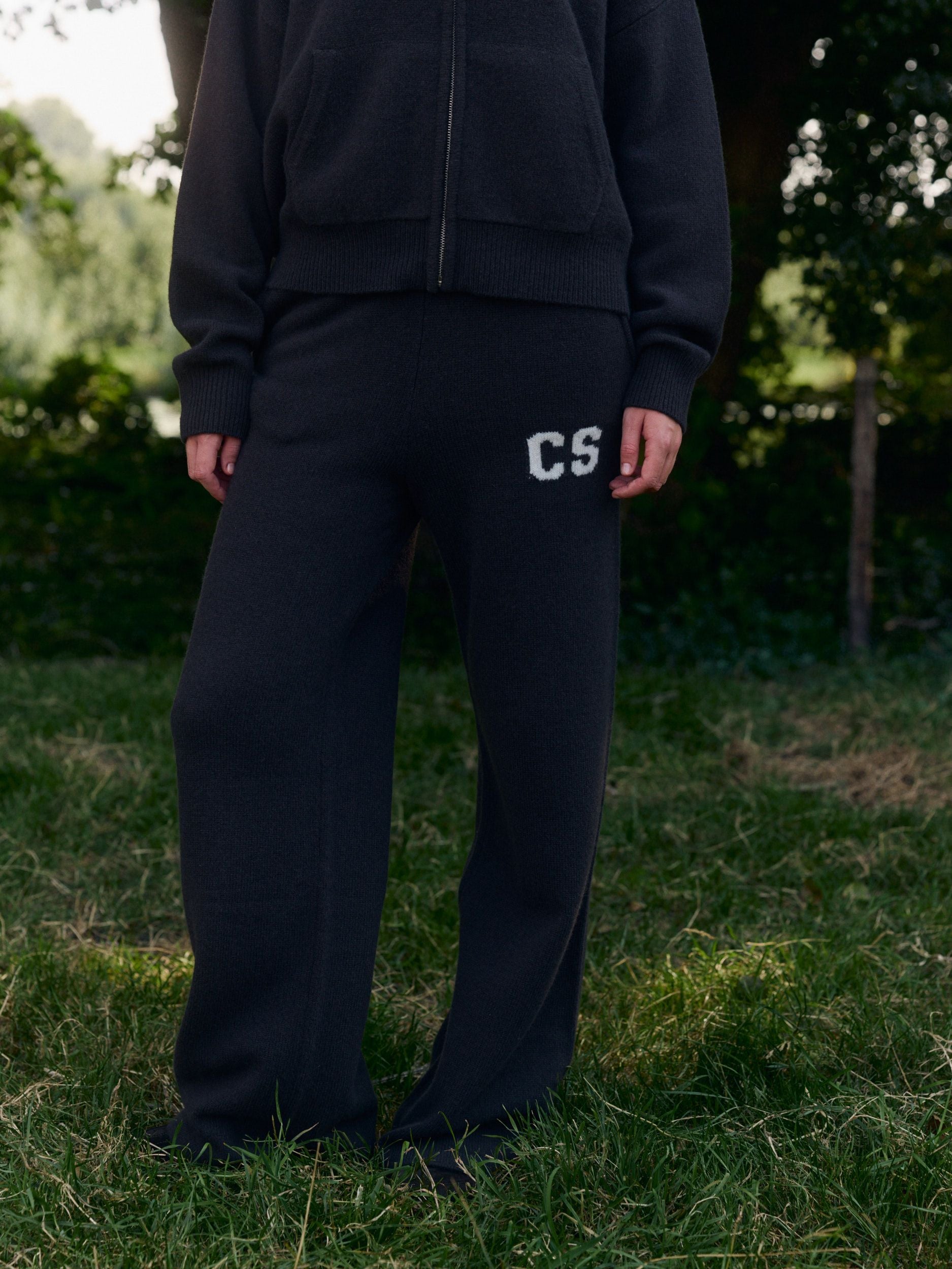 Woman wearing dark brown knit straight-leg pants with a matching zip sweater standing outdoors on grass.