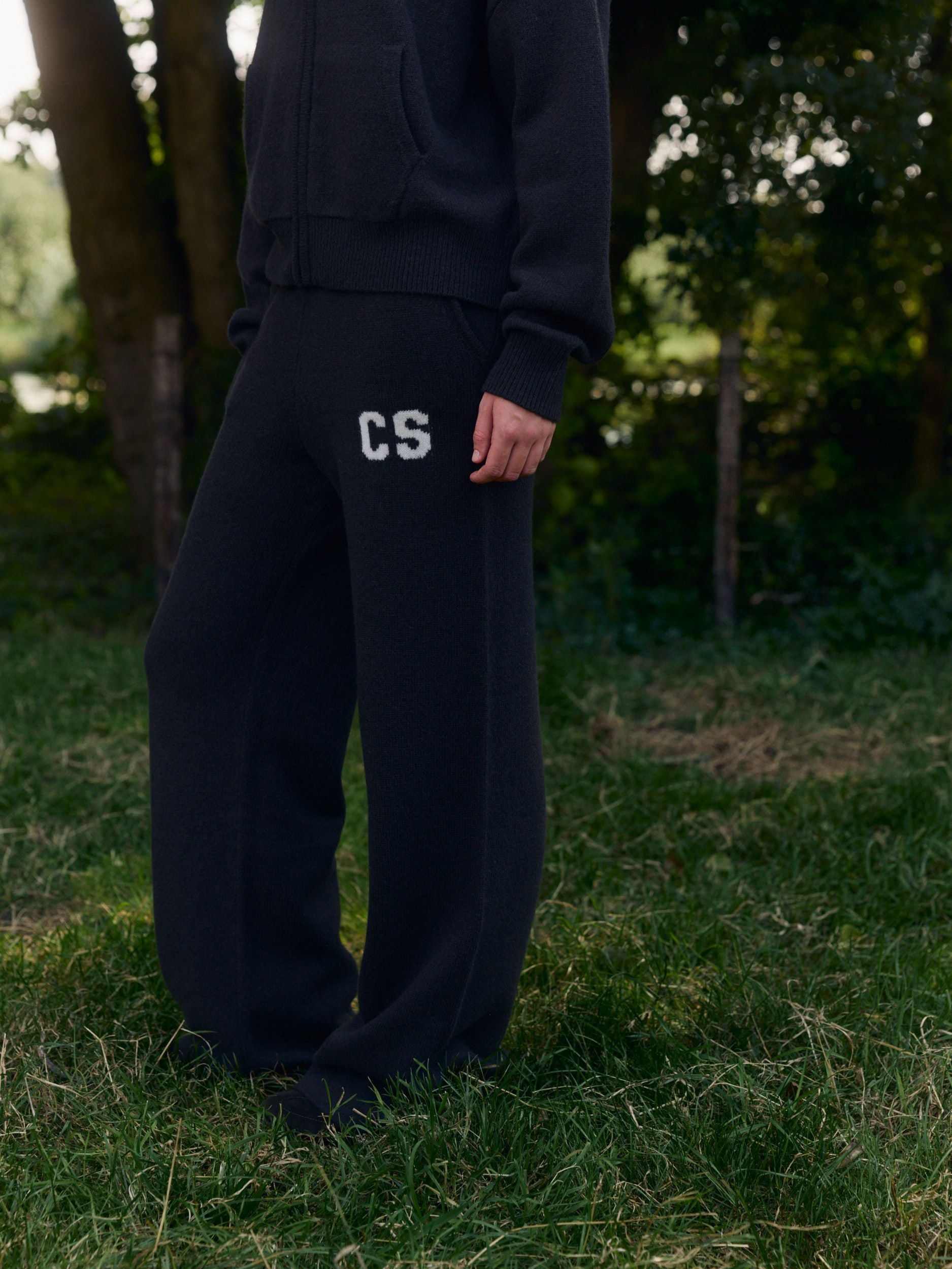 Woman wearing dark knit straight-leg pants with a CS logo standing outdoors on grass.