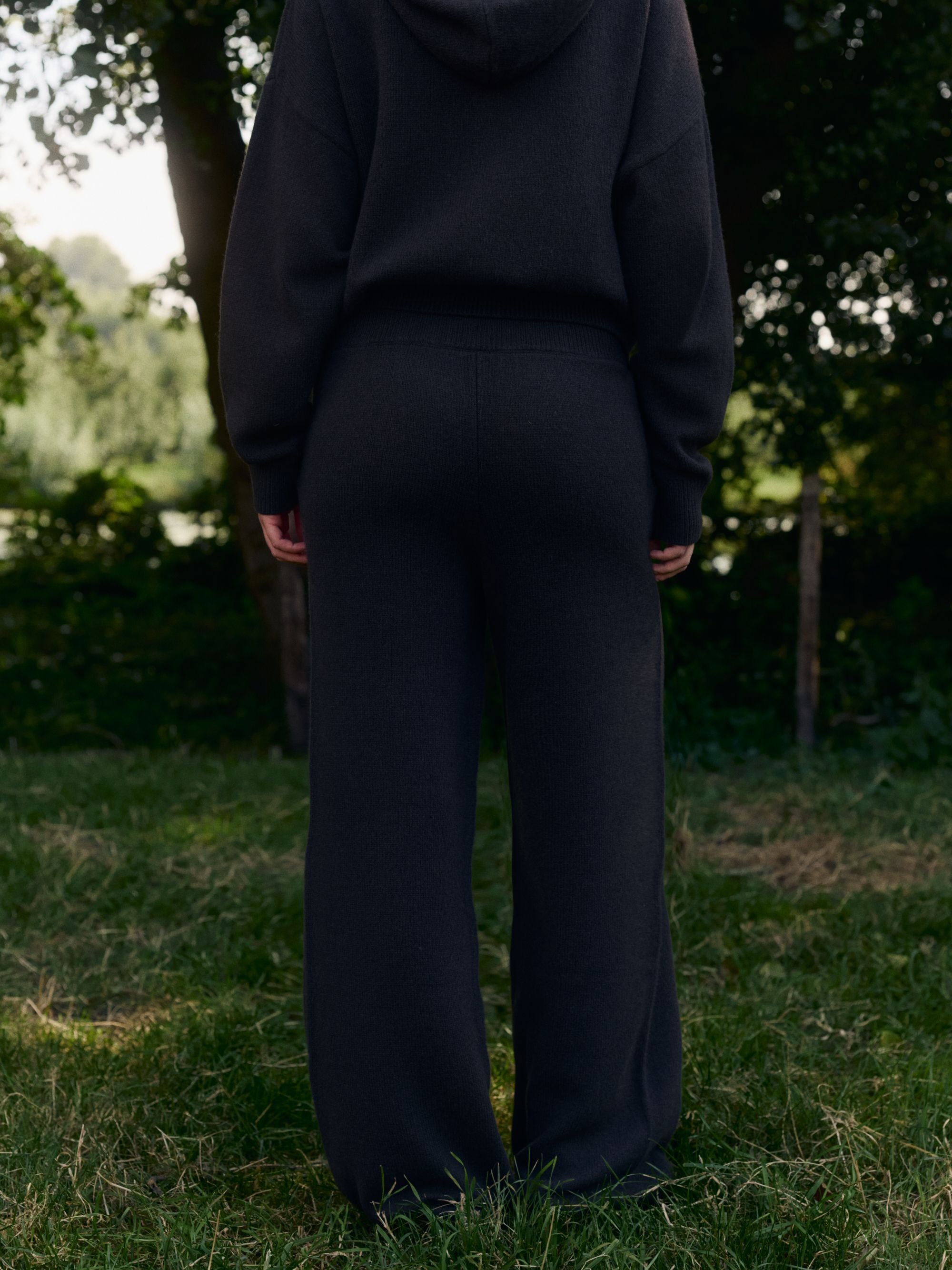 Woman wearing cocoa bean CASPARA woolknit straight-leg pants and matching hoodie outdoors, seen from behind.