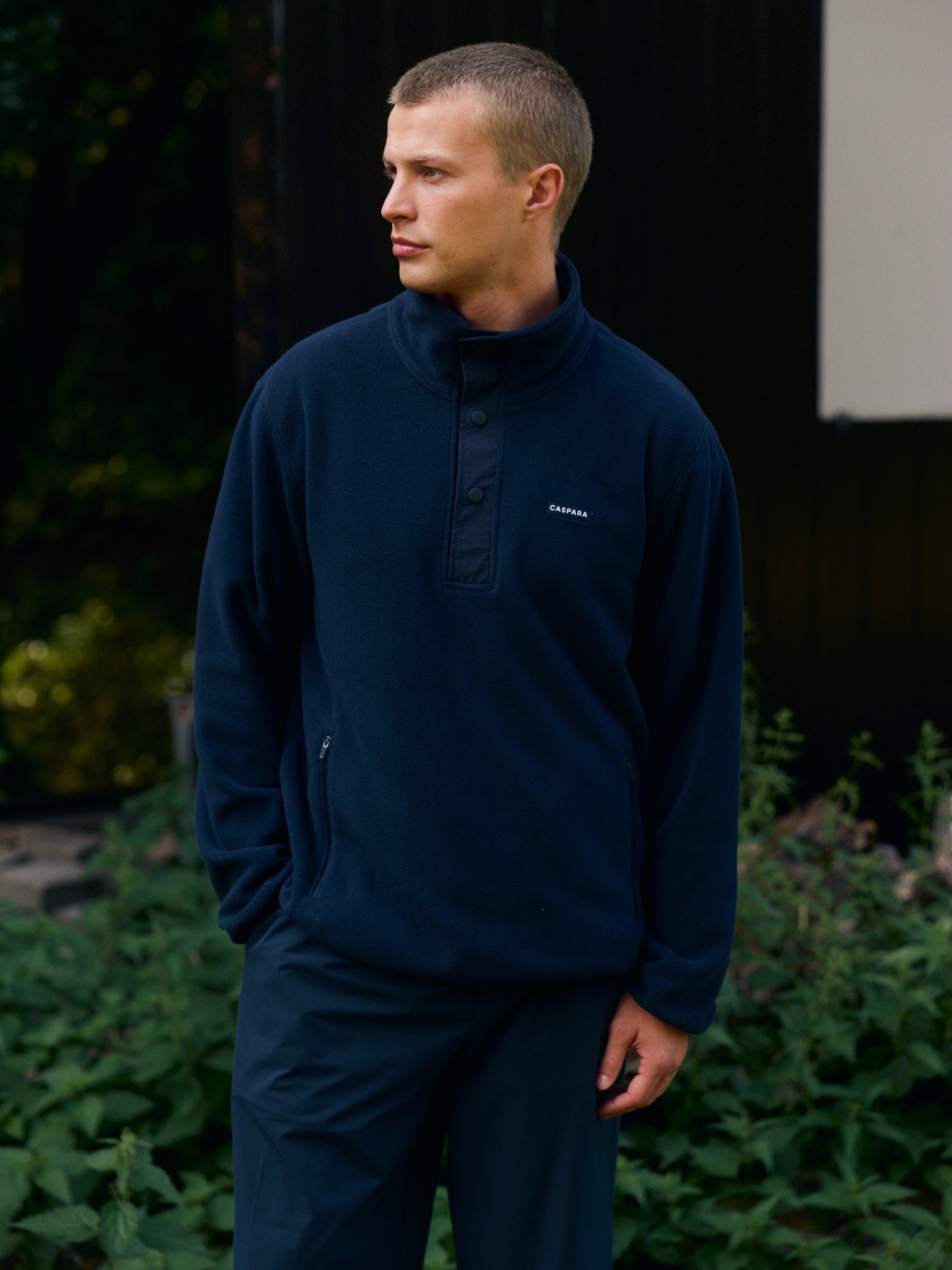 Man wearing a navy polar fleece half-snap pullover standing outdoors in front of greenery.