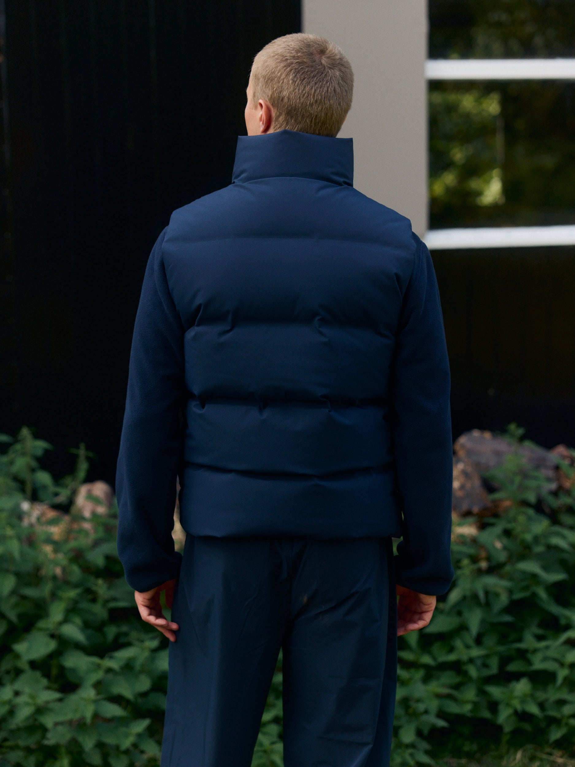 Back view of a man wearing a navy blue puffer vest layered over a fleece jacket outdoors.