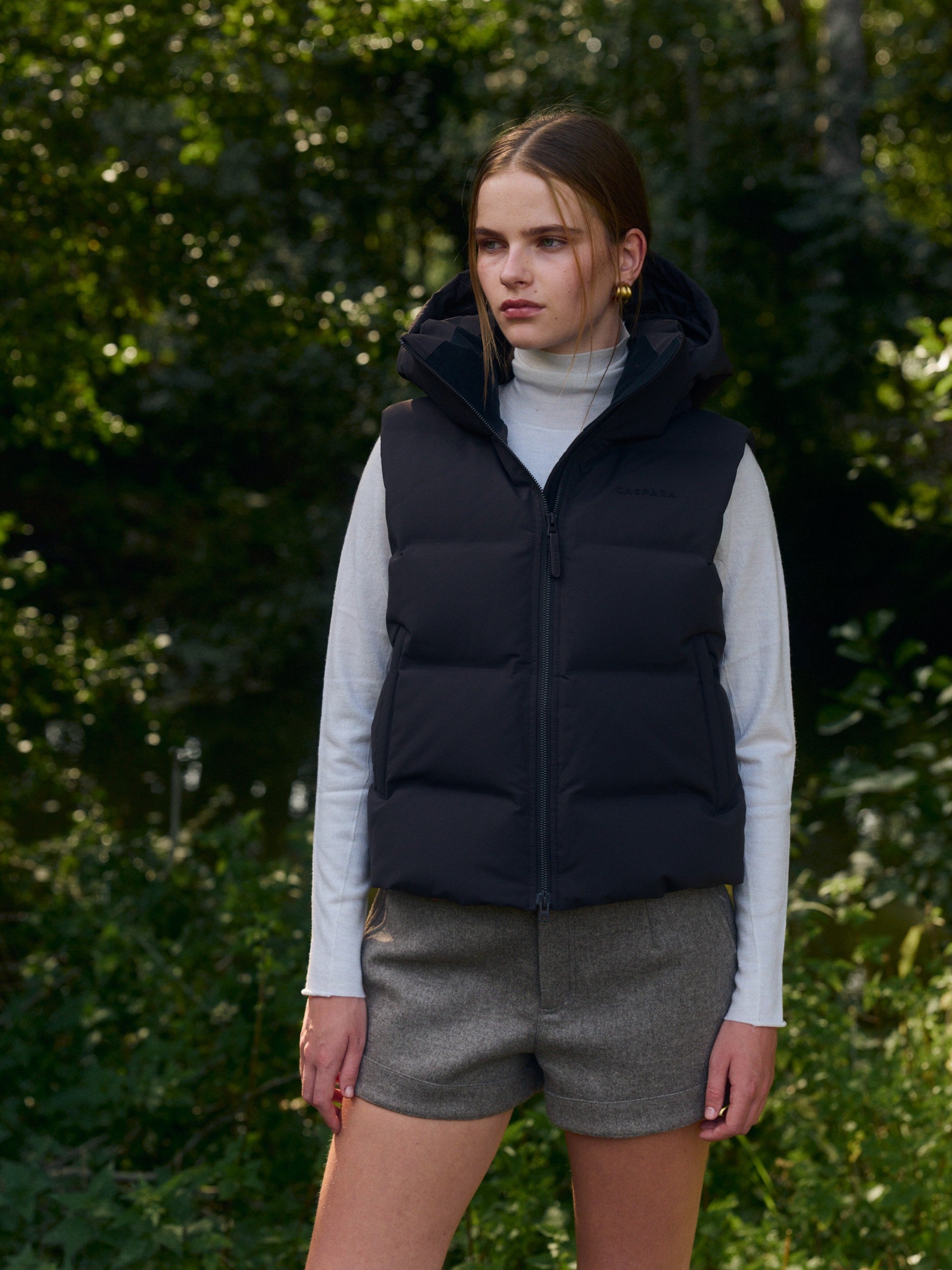 Woman wearing an espresso hooded puffer vest over a light turtleneck outdoors in a lush green setting.