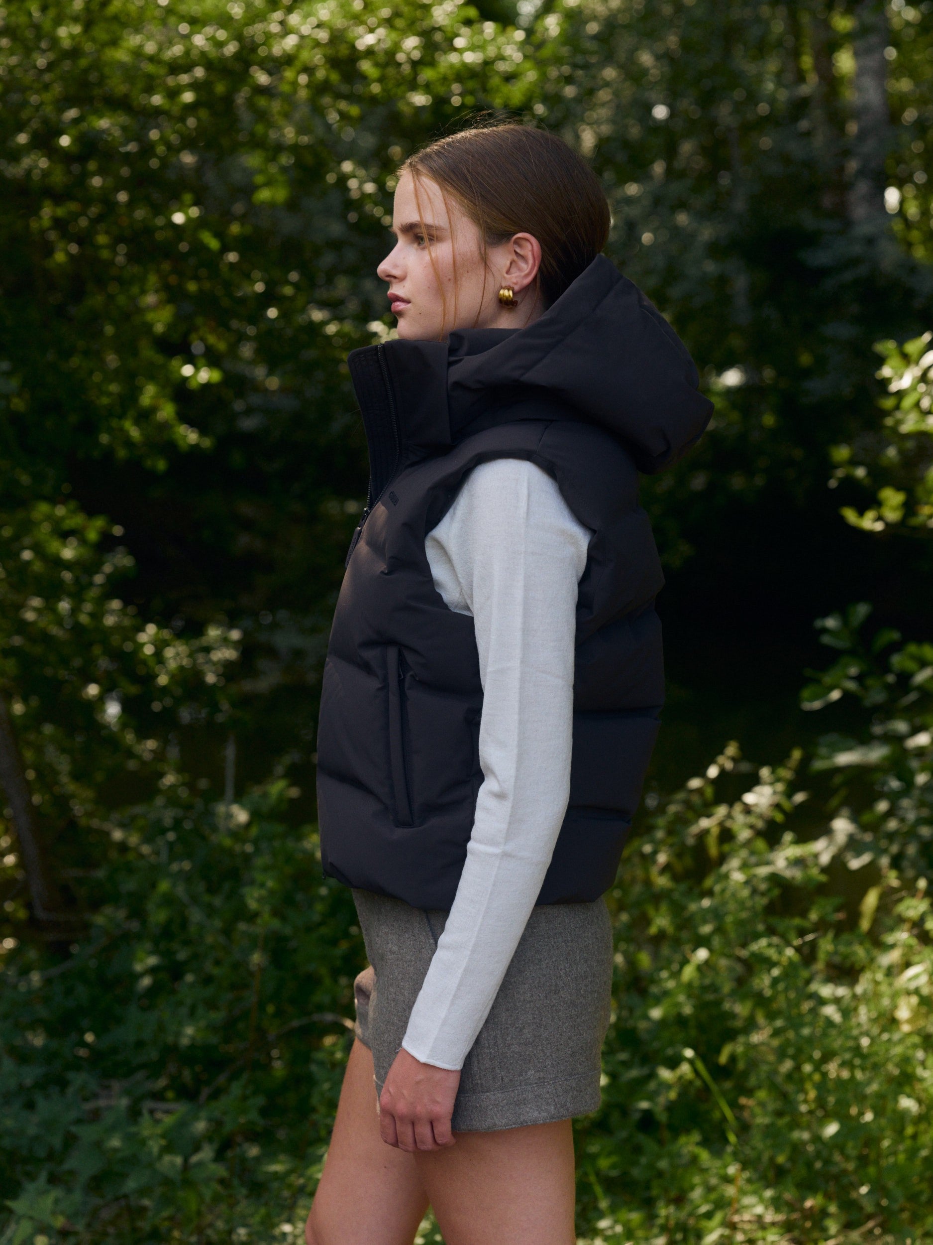 Woman wearing an espresso hooded puffer vest outdoors against lush green trees.