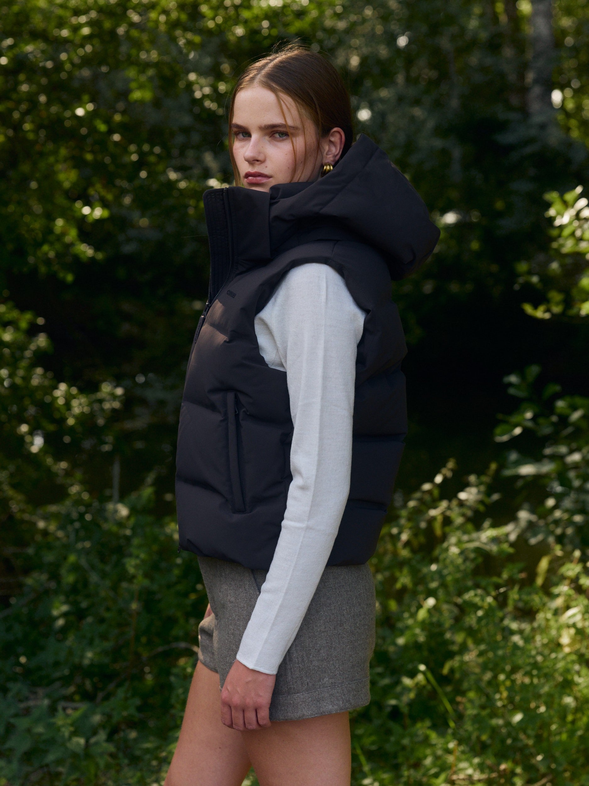 Woman wearing an espresso hooded puffer vest layered over a white long-sleeve top outdoors.