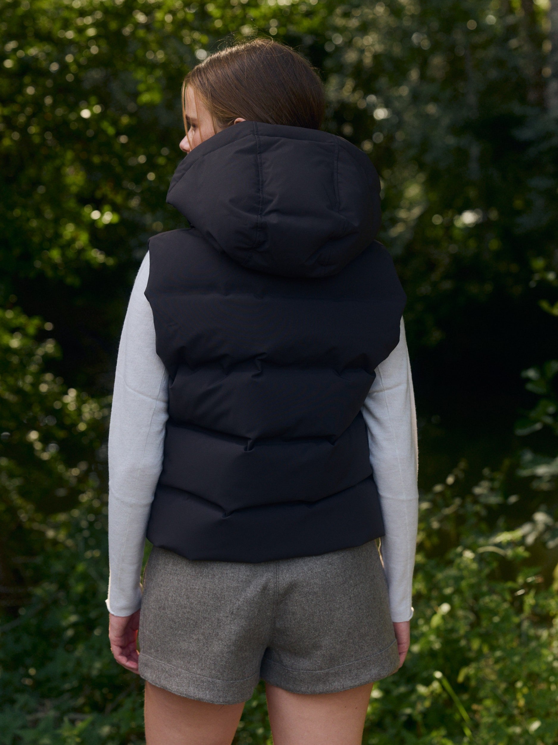 Back view of a woman wearing an espresso hooded puffer vest in an outdoor nature setting.