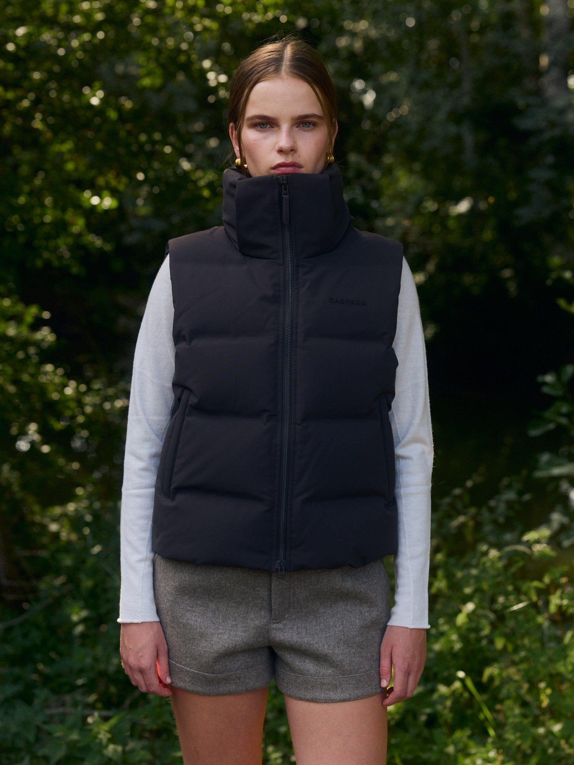 Woman wearing an espresso puffer vest with a high collar standing outdoors in front of trees.