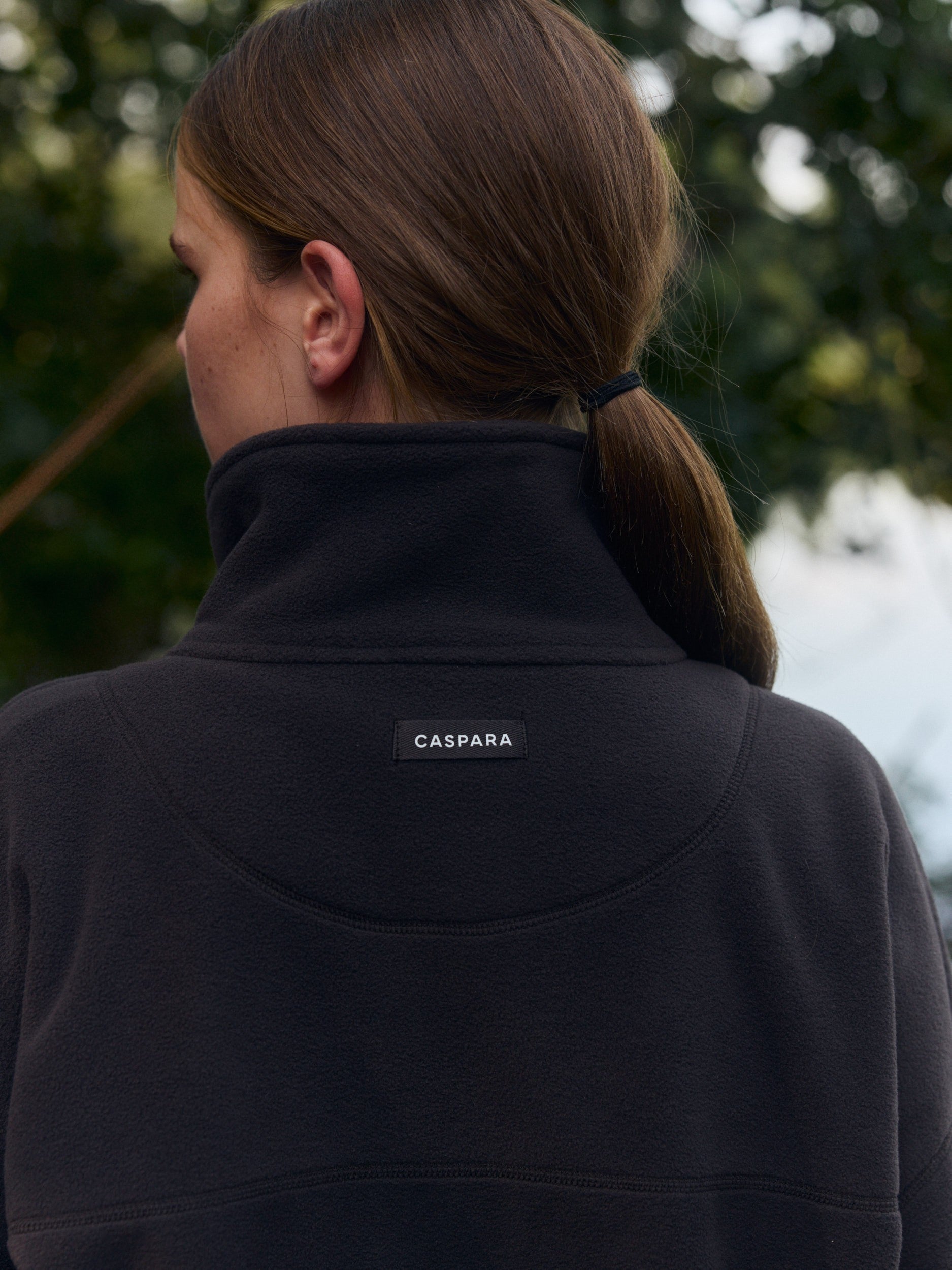 Back view of a woman wearing an espresso polar fleece pullover with a high collar and logo patch.