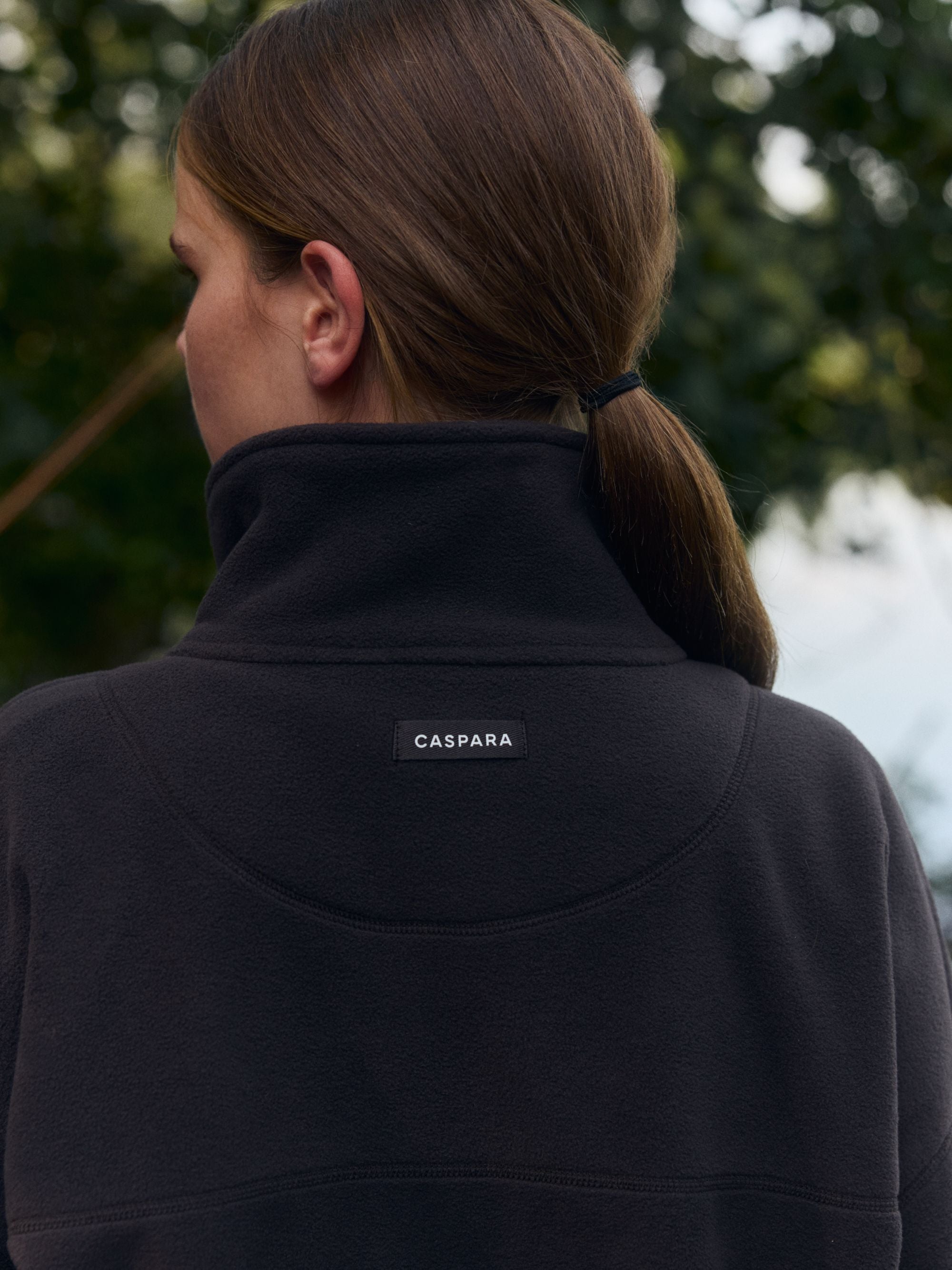 Back view of a woman wearing an espresso CASPARA polar fleece pullover outdoors.