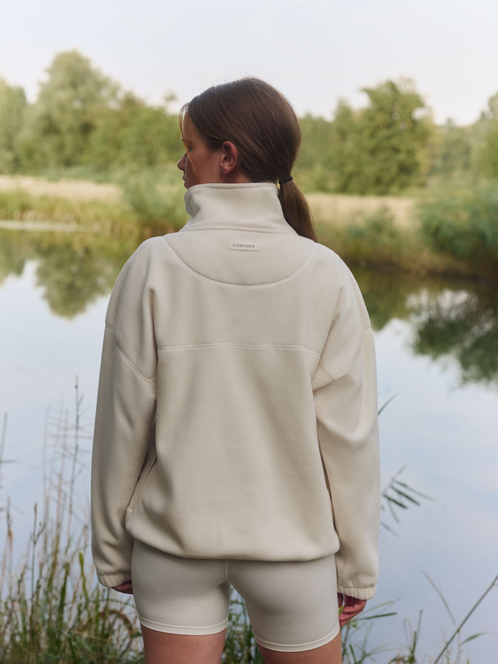 Back view of a woman wearing a creme CASPARA polar fleece pullover outdoors by a lake.