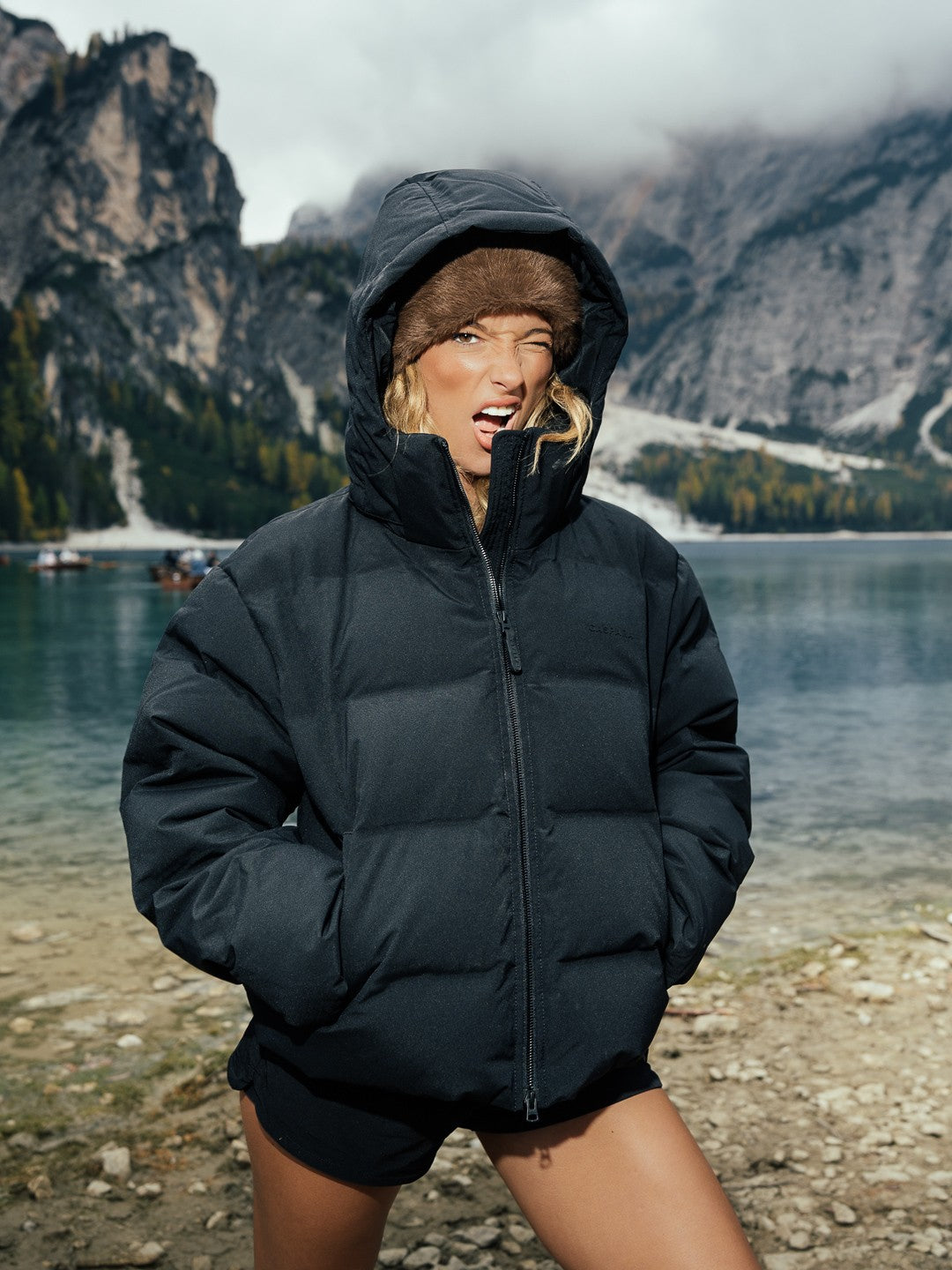 Woman wearing a black CASPARA Aurora puffer jacket with the hood up by an alpine lake.