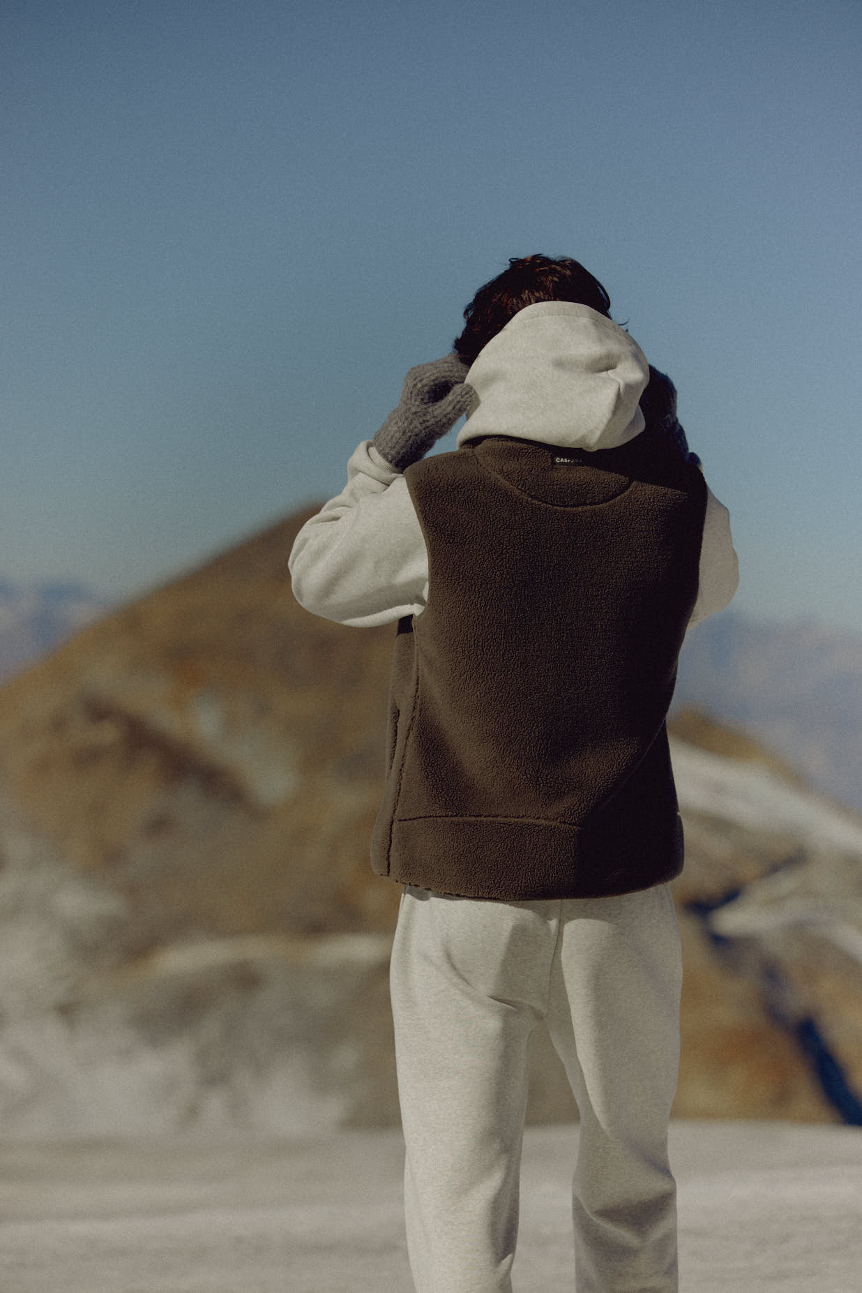 Rear view of a man wearing a khaki CASPARA Cloudfleece vest in a mountain landscape.