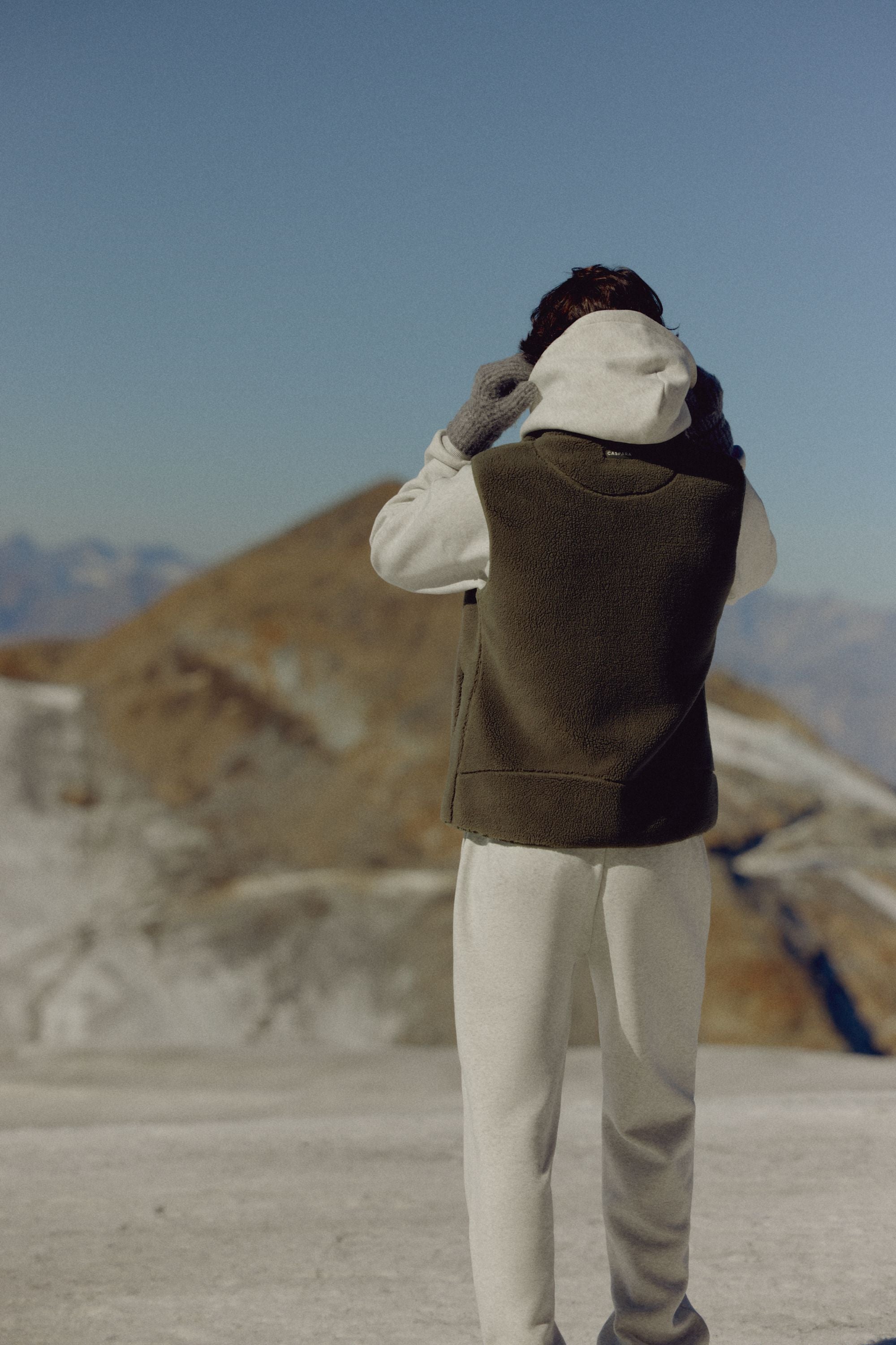 Back view of a man wearing a khaki CASPARA Cloudfleece vest in a mountain landscape.