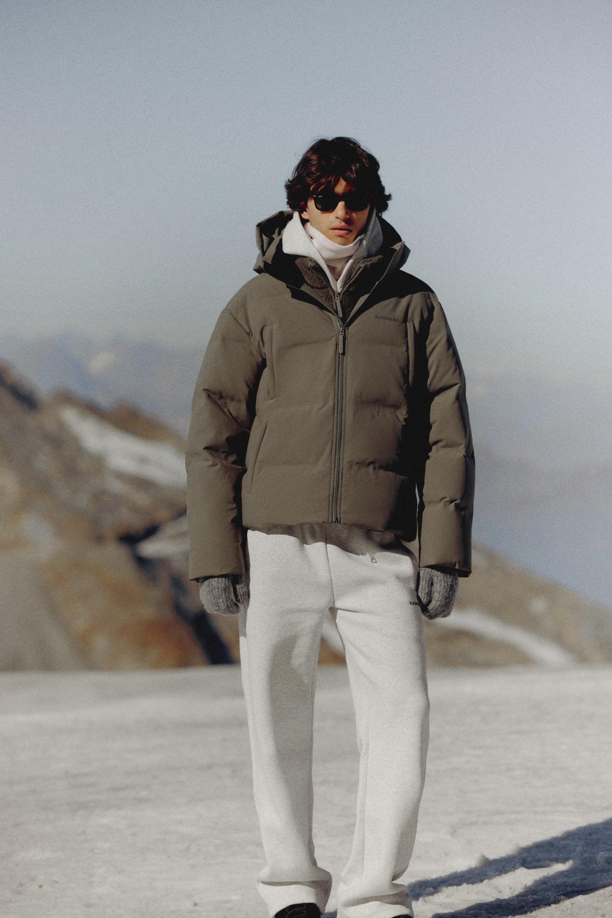 Man wearing a khaki CASPARA Boreal puffer jacket in a snowy mountain landscape.