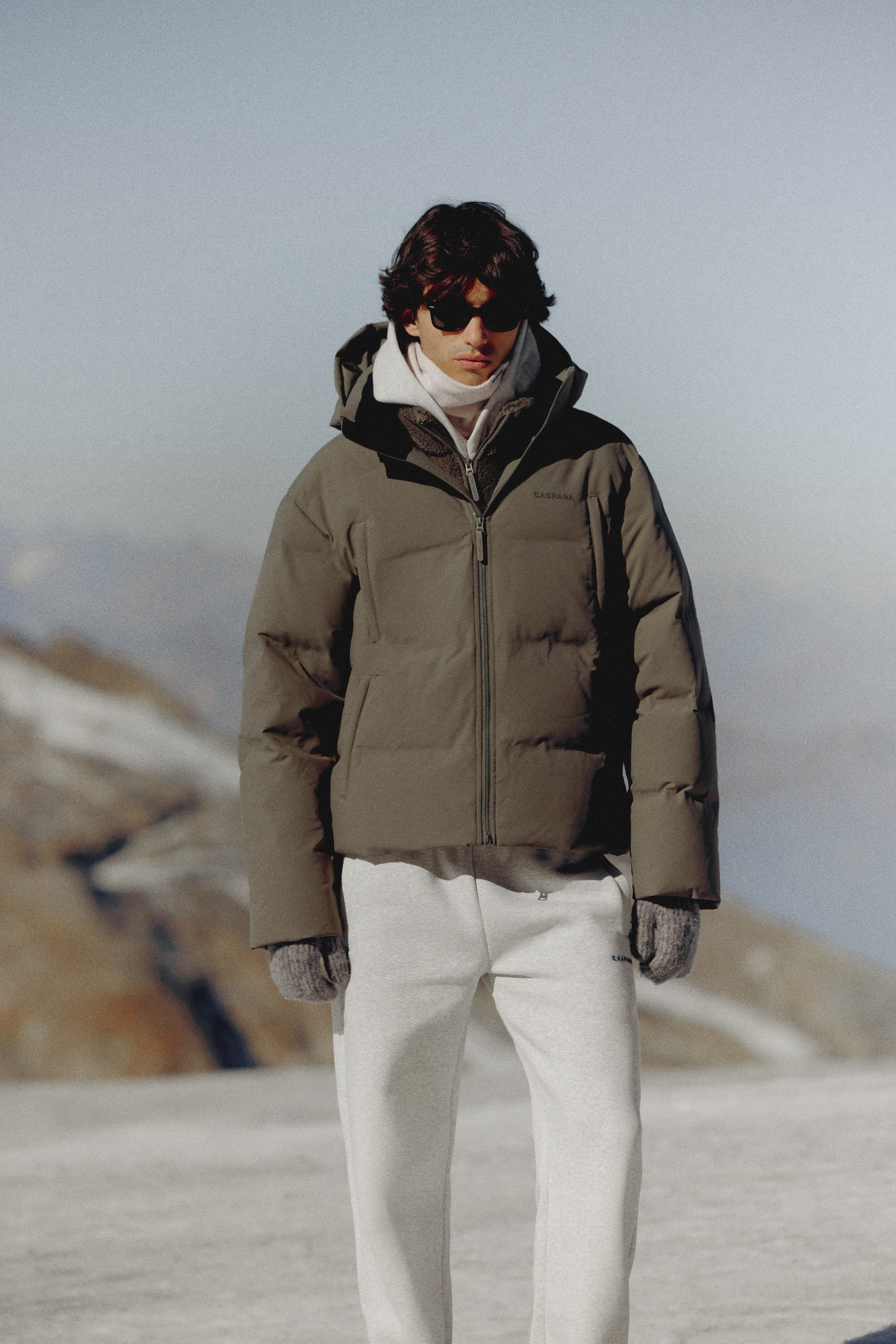 Man wearing a khaki CASPARA Boreal puffer jacket in a snowy mountain setting.
