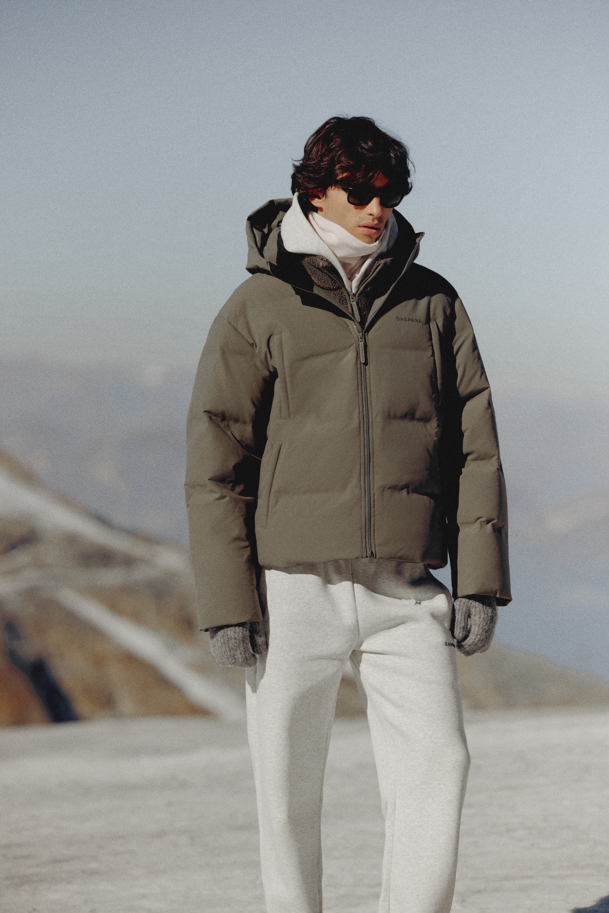 Man wearing a khaki CASPARA Boreal puffer jacket in a snowy mountain setting.